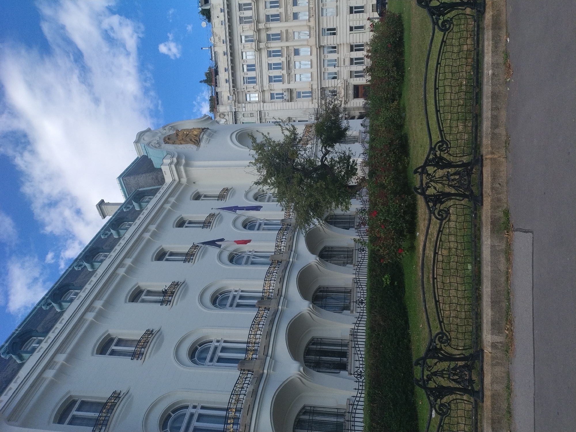 The image depicts the French Embassy in Vienna, Austria, situated on a tree-lined street. The building is a grand white structure with multiple floors, featuring arched windows on the lower levels and rectangular windows on the upper floors. A balcony with an ornate black and gold railing runs along the first floor. French flags are visible on the building. To the right of the French Embassy, another large white building with a more uniform facade and numerous windows is visible. In the foreground, a black wrought-iron fence separates the street from a manicured lawn and a dense hedge with red roses. The sky is blue with scattered white clouds. The scene is captured from a low angle, looking up at the buildings.