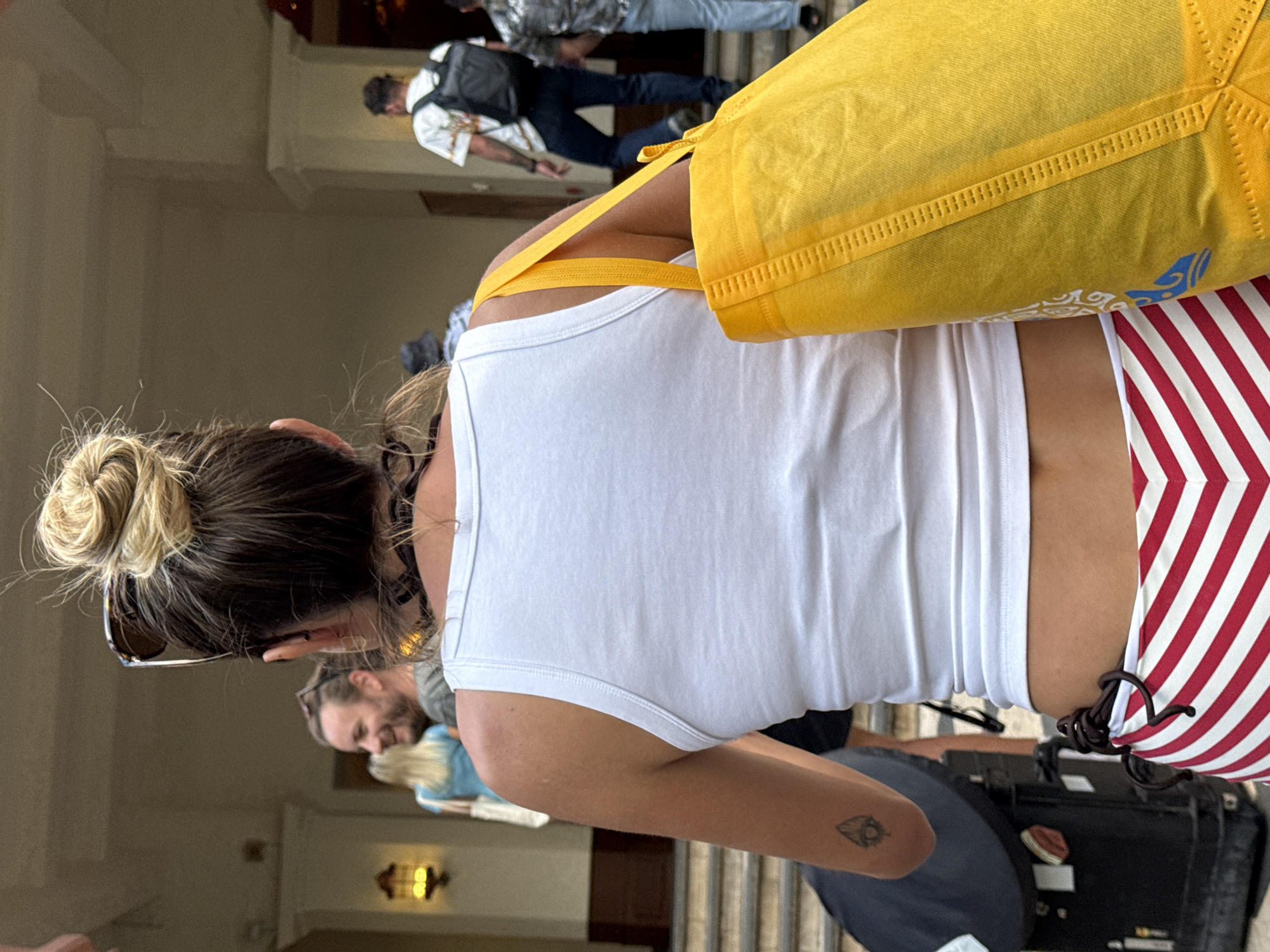The photograph shows a woman from the back wearing a white tank top, a striped bathing suit bottom, and a yellow bag. She has brown hair in a bun. Sunglasses are perched on top of her head. The woman has a tattoo on her right arm. In the background, there are other people, stairs, and a building. The scene is located in Honolulu, United States of America.