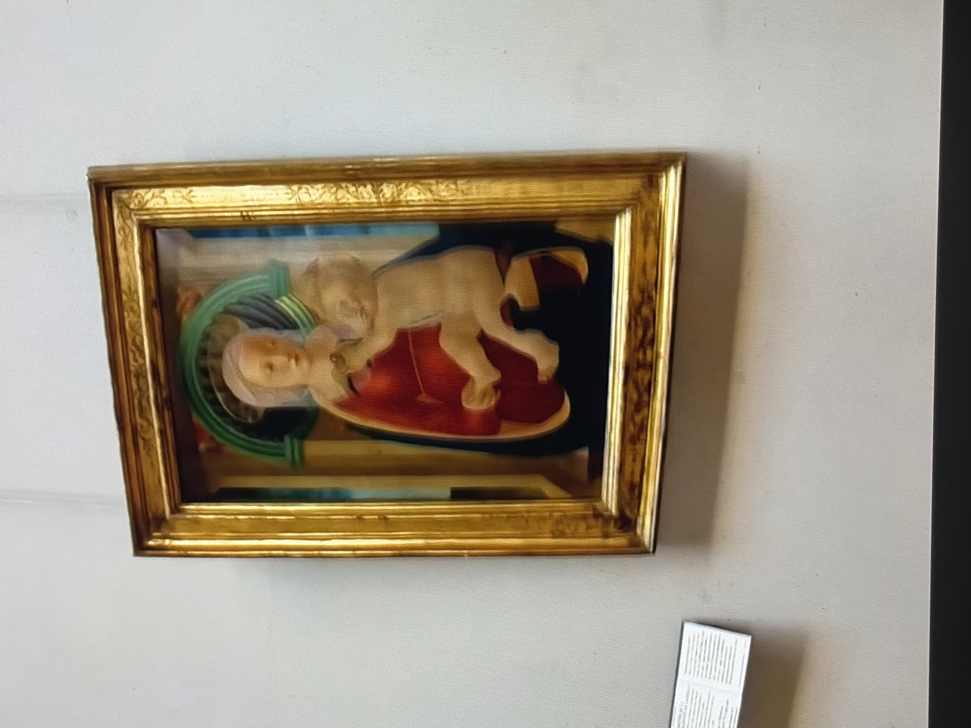 An oil painting of the Madonna and Child, framed in gold, hangs on a white wall. The painting depicts the Virgin Mary in a red robe and headscarf, holding a nude infant Jesus. The background is blue with an arched green element. A small informational plaque is to the left of the painting.
