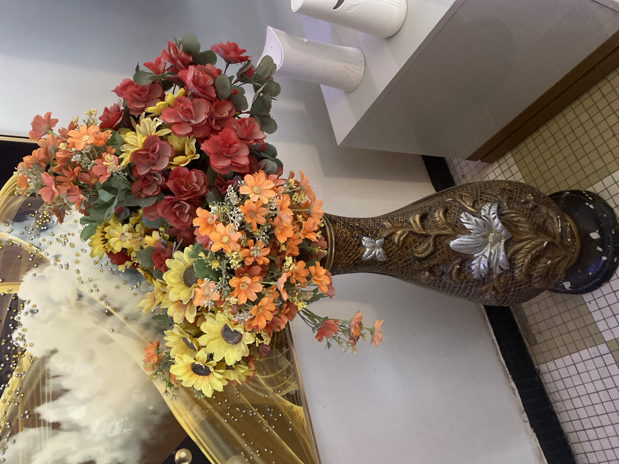 A tall, ornate vase filled with a vibrant bouquet of artificial flowers sits on a tiled floor. The vase is predominantly brown with a textured, woven pattern and decorated with metallic silver floral accents. The flowers in the vase are a mix of red, orange, and yellow hues, including some sunflowers and smaller daisy-like blooms. They are arranged in a full, cascading manner, with some greenery interspersed. To the left of the vase, a decorative element with a glossy, golden surface and scattered embellishments is visible, reflecting the light and surrounding environment. In the background, a white wall meets a light gray surface, possibly a shelf or ledge, upon which two white cylindrical objects are placed. The floor is covered in small, square tiles in a cream and beige checkered pattern. The overall scene suggests an indoor setting, perhaps a home or a commercial space, decorated with floral arrangements. The lighting appears to be artificial, as there are no clear indications of natural daylight. No people are visible in the frame, and no text is discernable. The arrangement of the flowers and the decorative vase create a visually appealing display.