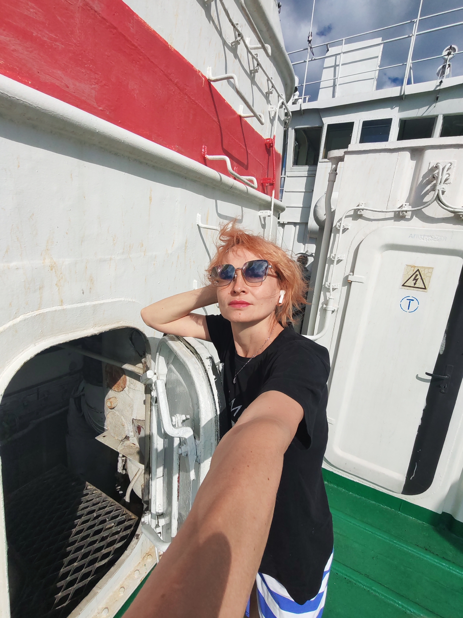 A medium shot, taken from a low angle, captures a woman with short, orange hair and sunglasses, wearing a black t-shirt and striped shorts, leaning against the white hull of a ship. She holds one arm up, resting her head on her hand, and looks towards the camera. The ship's hull is painted white with a thick red stripe running horizontally near the top. To the right of the woman is a white door or hatch with an electrical hazard symbol and a blue circular symbol. The deck is painted green. The background shows the white superstructure of the ship and a cloudy blue sky. The image appears to have been taken on a sunny day. The location is identified as Krokhino, Russia, a place known for its abandoned church on the bank of the Sheksna River.