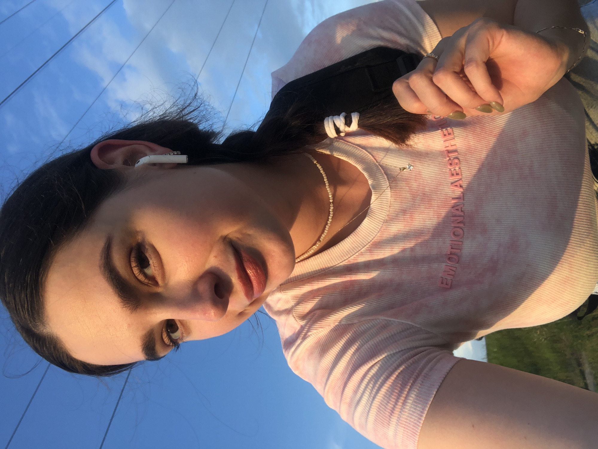 This is a medium-shot, vertical, selfie-style photograph taken outdoors, likely during the golden hour due to the warm lighting. The subject is a young woman with dark hair, which is partially pulled back, and she is wearing white earbuds in her right ear, adorned with small rhinestones. She has on a tie-dye t-shirt with pink and white coloring, and a subtle necklace. The text "EMOTIONAL AESTHETICS" is visible in pink lettering on her shirt. Her fingernails are painted a muted olive green, and she is wearing a ring on her right ring finger. Her expression appears relaxed and content. The background consists of a bright blue sky with wispy clouds and horizontal power lines stretching across the frame, suggesting an open, possibly rural or suburban, outdoor setting. The lower portion of the image reveals a hint of green grass, further supporting an outdoor location. The overall mood is casual and serene.