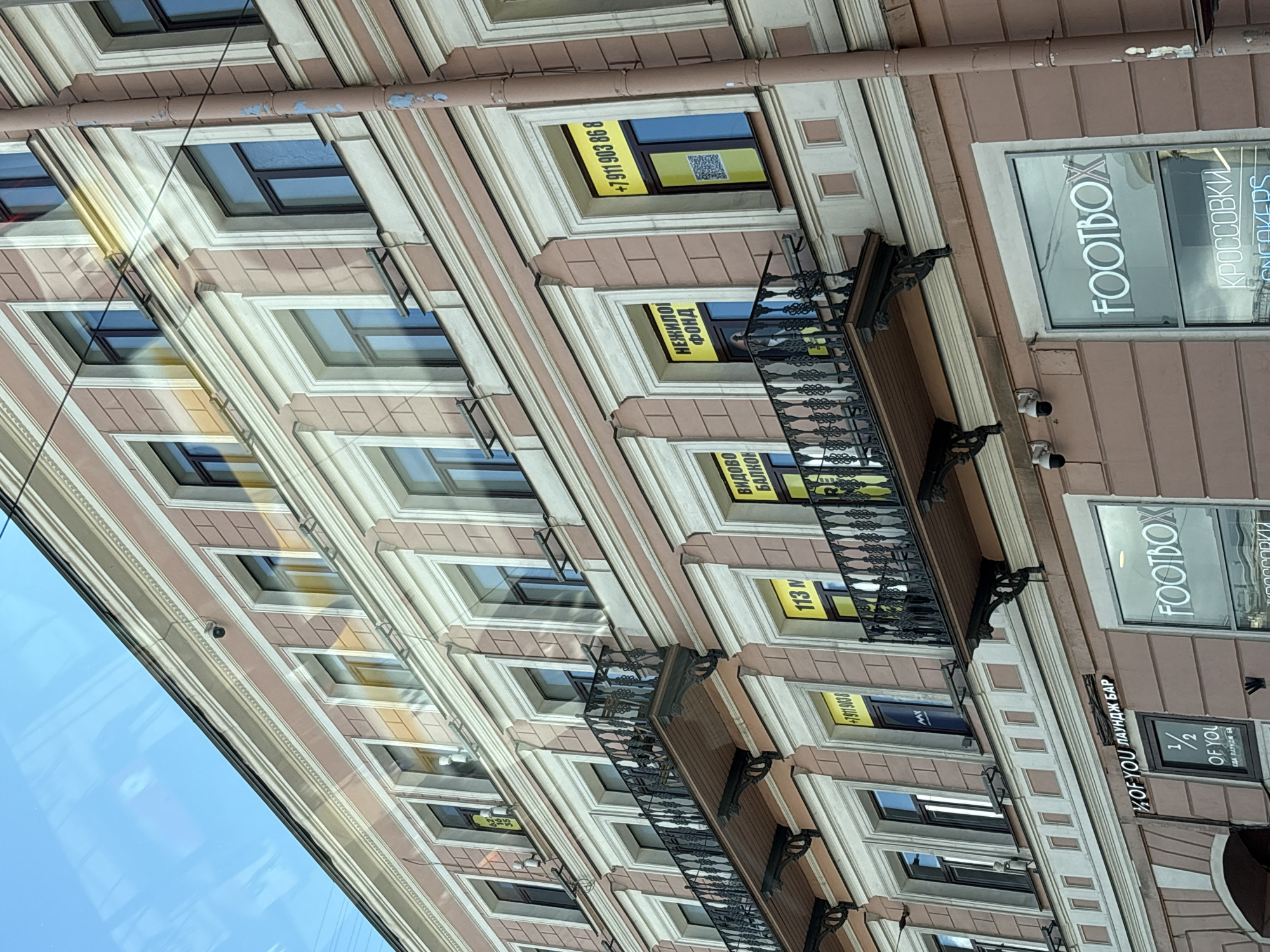 The image shows the facade of a multi-story building in Saint Petersburg, Russia. The building has a pinkish-brown color with white decorative elements around the windows and along the cornices. Several windows are visible, some with black frames, and some with reflective glass showing the sky and surrounding environment.
On the upper floors, there are several balconies with black wrought-iron railings. Some windows have yellow signs with black text. One sign reads "+7 911 903 86 8", another "НЕЖИЛОИ ФОНД", and another "ВИДОВО БАЛКОН". There are also some security cameras mounted on the building.
On the lower level of the building, there are shopfronts. One shop has a sign that reads "FOOTBOX" in large white letters. Below this sign, the word "КРОССОВКИ" (Sneakers) is visible. Another shopfront has a sign that says "1/2 OF YOU" and "ЕДА ЛАУНДЖ БАР".
The overall impression is of a street view of a commercial building in a city. The lighting suggests it is daytime, with sunlight reflecting off the windows, creating some lens flares and glare.