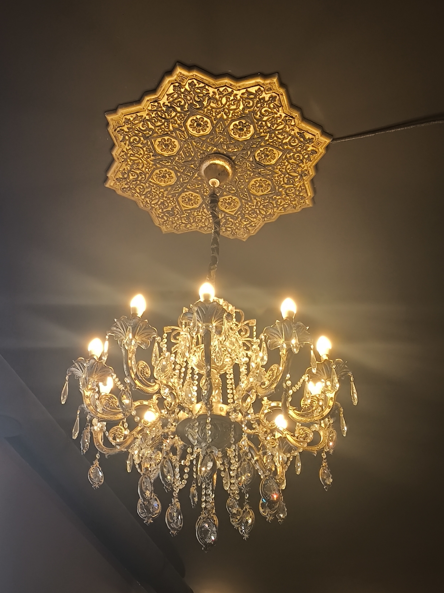 This image captures an ornate, vintage-style chandelier hanging from a ceiling adorned with a decorative gold medallion. The chandelier is made of gold-toned metal and features numerous crystal droplets that reflect the warm light emitted by its glowing bulbs. The ceiling medallion is intricately carved with geometric and floral patterns, suggesting a traditional or classical architectural style. The background is a dark, neutral color, likely a ceiling, creating a dramatic contrast that highlights the illuminated chandelier and medallion. The lighting is soft and warm, with visible rays of light emanating from the bulbs, adding a sense of ambiance and luxury. The overall impression is one of elegance and opulence. There is no visible text in the image. The setting appears to be indoors, possibly in a room with high ceilings, such as a ballroom, dining room, or a grand hall. The image conveys a sense of sophisticated decor.