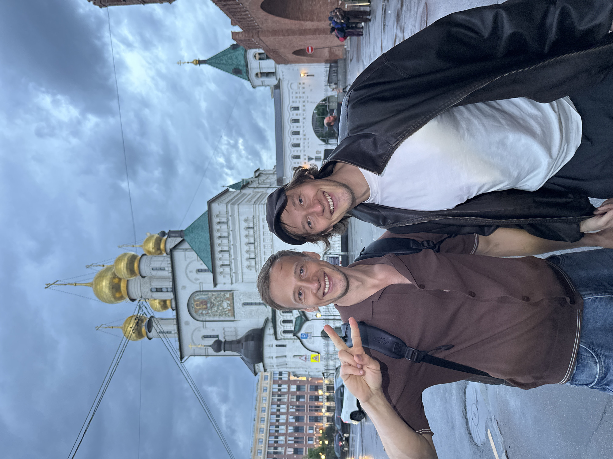 Two men pose for a photo in front of the Church of the Transfiguration, a historic Russian Orthodox church located in the historic center of Saint Petersburg, Russia, near the Moscow railway station. The sky is overcast, and the ground appears wet, suggesting recent rain. The man on the left, wearing a brown polo shirt and blue jeans, smiles and holds up a peace sign. He has a backpack on. The man on the right, wearing a white t-shirt, black leather jacket, and a black baseball cap, also smiles. In the background, other people and cars are visible. The architecture of the church, with its golden domes and intricate detailing, is prominent. The overall atmosphere is subdued due to the weather and time of day.