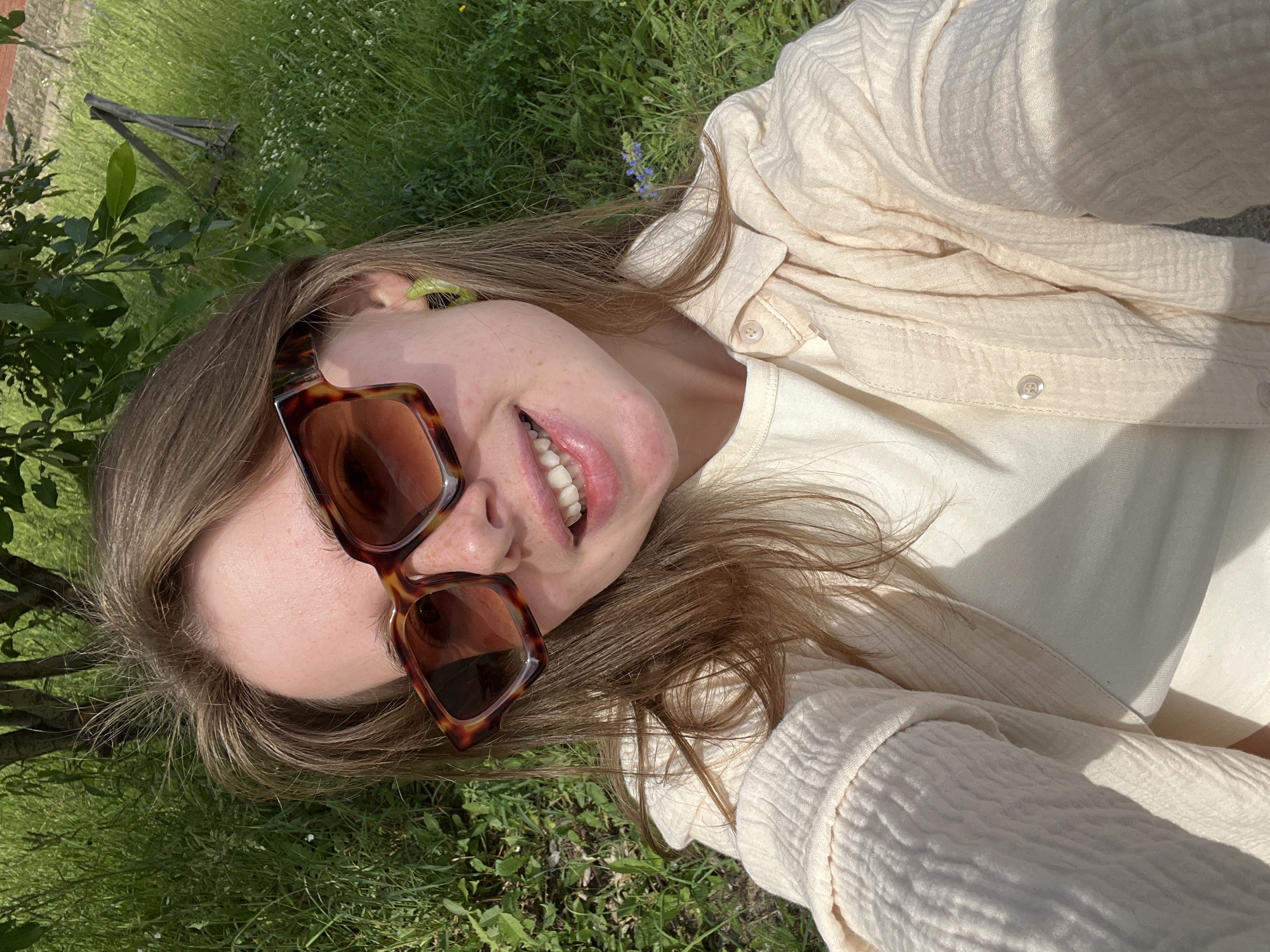 A young Caucasian female is captured in a selfie outdoors, wearing oversized tortoiseshell sunglasses and a light-colored collared shirt over a cream-colored top. Her hair is light brown and falls loosely around her shoulders. She has bright green hoop earrings. The background consists of lush green grass, trees, and a wooden fence post, suggesting a rural or park-like setting, possibly in the city of Chaikovsky, Russia, given the context. The lighting indicates it is daytime, with sunlight casting shadows.