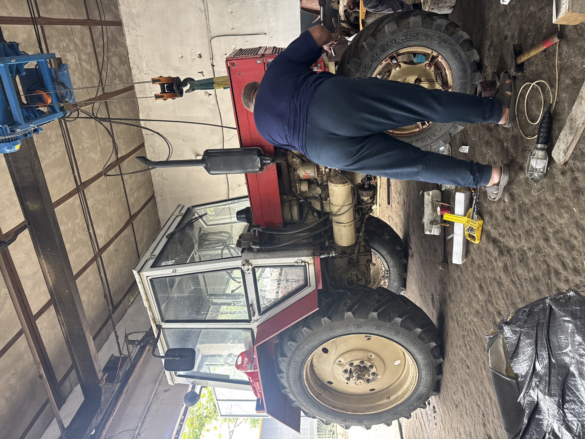 A man is working on a red and white tractor in what appears to be an indoor workshop or garage in Sarnevo, Bulgaria. The image is rotated 90 degrees clockwise, showing the man from behind, leaning over the engine bay of the tractor. He is wearing a dark blue long-sleeved shirt and matching pants, with sandals on his feet.
The tractor, likely a utility or agricultural model, has a red body, a white-framed cabin with glass windows, and large cream-colored wheels. The front right tire clearly shows "VOLTYRE" and "D-220 M-35" text. The engine compartment is open, revealing various mechanical components, hoses, and an exhaust pipe extending upwards.
Above the tractor, a blue industrial hoist or lifting mechanism is suspended from the ceiling structure, with cables leading down to a hook assembly. This suggests heavy components might be moved or have been moved recently during the repair. The ceiling consists of visible metal beams and what appears to be corrugated or insulated panels.
The floor of the workshop is concrete and shows signs of wear and dirt, consistent with a working environment. Scattered on the floor near the tractor are tools and equipment, including a work light with a cage, a hammer with a wooden handle, and a yellow tool which might be a jack or clamp. A black tarp or cover is also visible on the floor to the right. The overall setting is functional and well-used, indicating ongoing maintenance or repair work on the tractor.