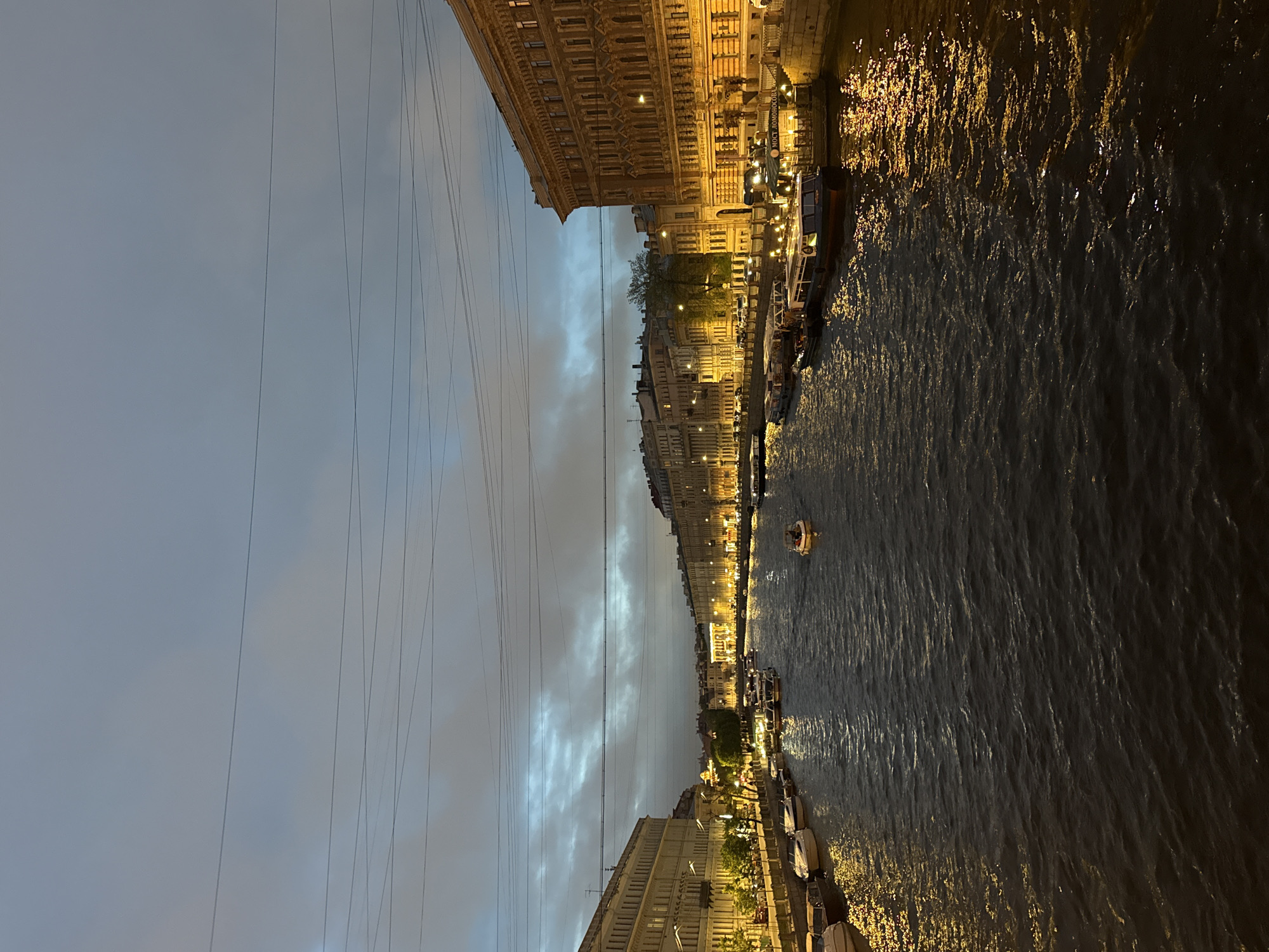 A nighttime scene shows a body of water reflecting city lights, bordered by buildings, under a cloudy sky. The water surface is rippled, reflecting the yellow lights from the buildings that line the banks. The buildings are European-style, with detailed facades. Several boats are docked along the banks, and one small boat is moving on the water. Above, power lines stretch across the sky, which is gray and overcast. The location is likely a canal in Saint Petersburg, Russia, due to the architectural style and abundance of canals in the city.