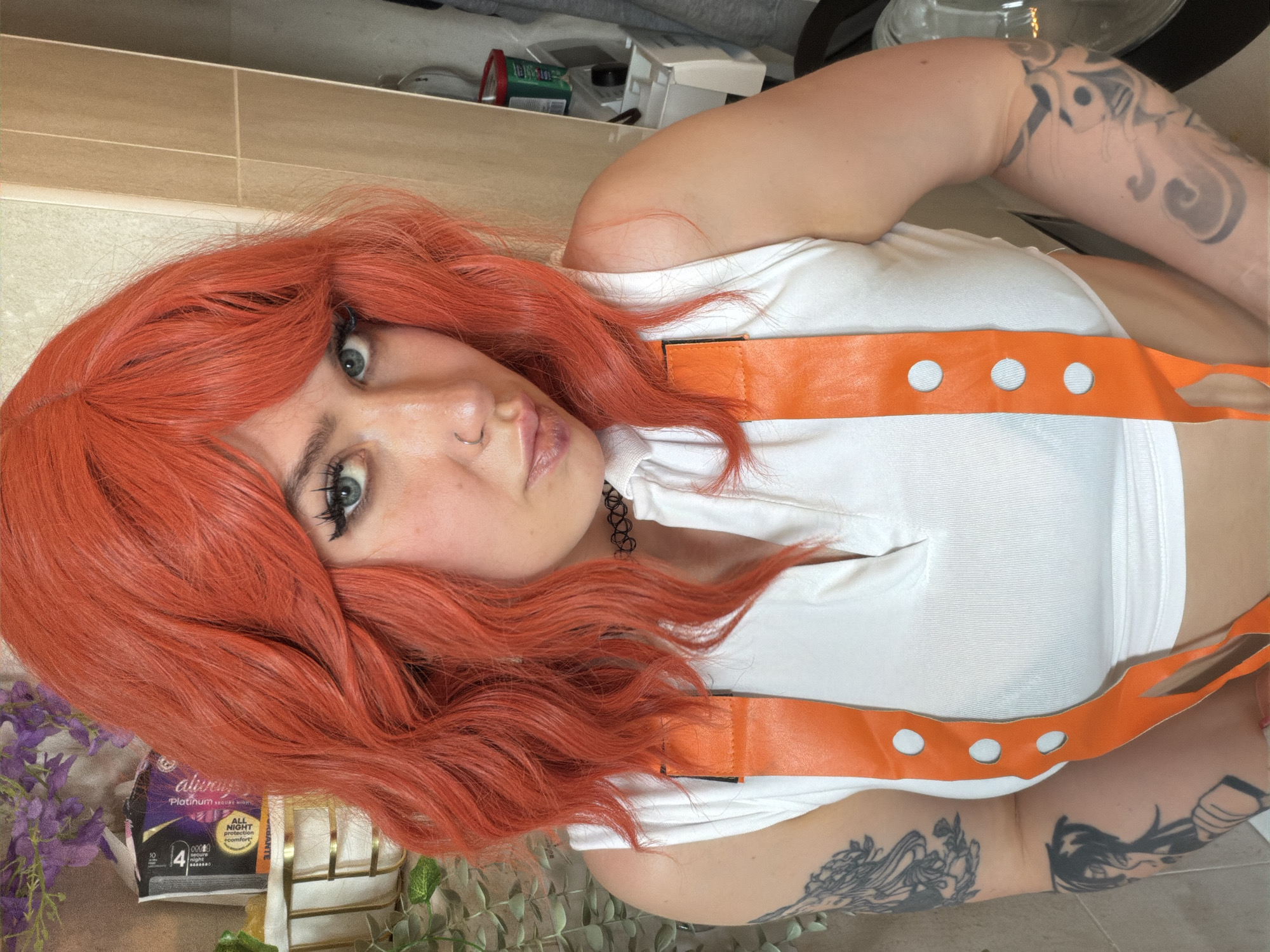 The image features a person with vibrant, shoulder-length, reddish-orange hair styled with a fringe and loose waves. The person, likely female, has striking blue-green eyes enhanced with dark eyeliner and mascara. A small silver hoop nose piercing is visible on their left nostril, and a black choker necklace is worn around the neck. They are wearing a white short-sleeved top with distinctive orange straps that have three circular cutouts on each side. Intricate black and grey tattoos are visible on both arms.
The person is posing for the camera, looking directly at the viewer with a slight pout. The image appears to be a selfie, taken at an angle, possibly from above.
The background shows an indoor setting, potentially a bathroom or dressing area, indicated by what looks like light-colored wall tiles or paneling. Various indistinct items, including a red-lidded container and a white box, are visible on a shelf or surface in the upper background. In the lower left corner, partially covered by the person's hair and some purple and green artificial plants, is a package of sanitary pads.
Visible text on the product packaging reads: "Always Platinum Secure Night," "ALL NIGHT protection comfort," "always," "4" (likely indicating a size), and "10 pads."
The general context for the image is Budva, Montenegro, though there are no specific visual cues within the image itself to confirm this location.