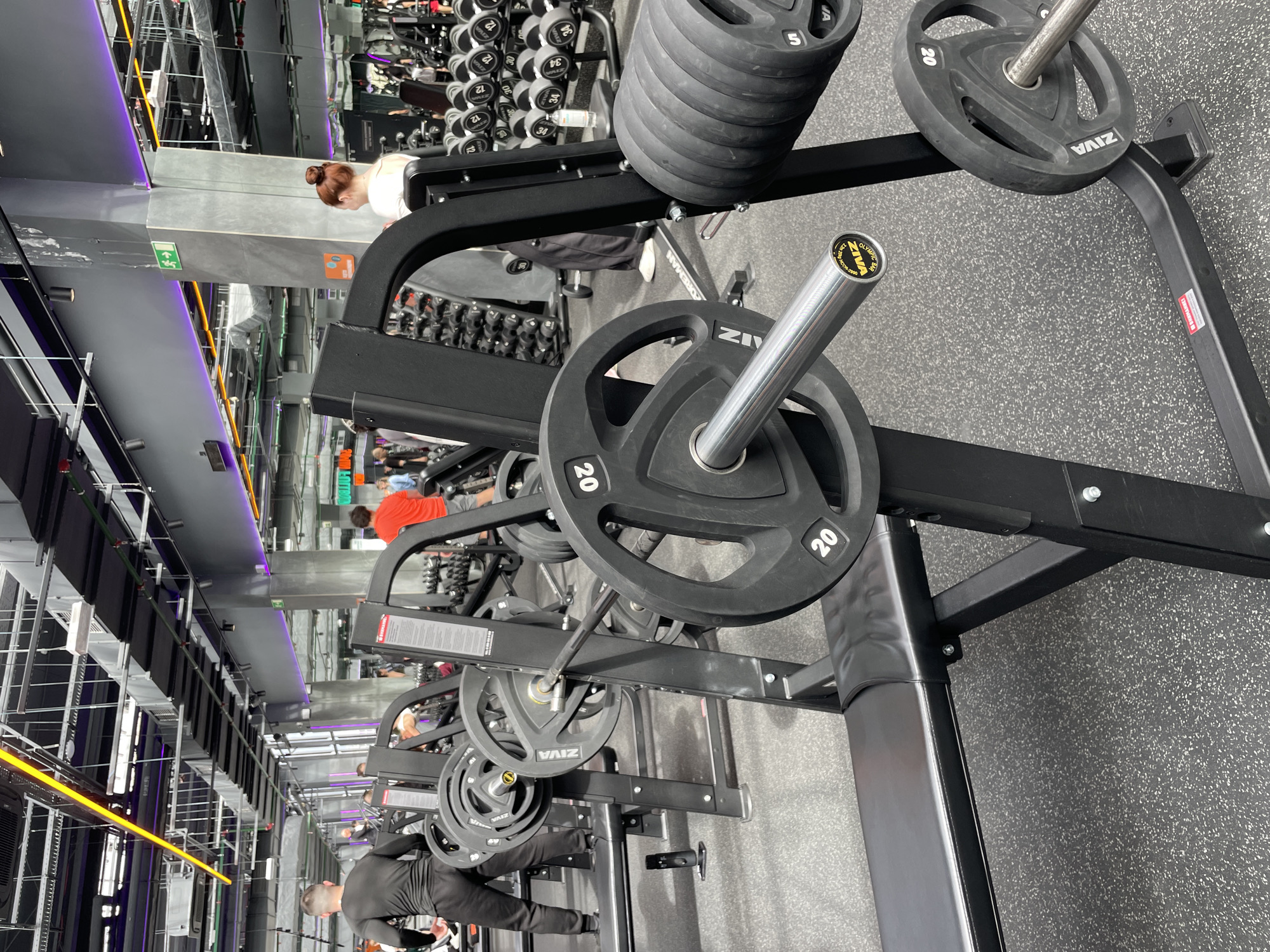 The image captures a workout area inside a gym. The foreground is dominated by gym equipment including weightlifting racks with barbells and weight plates. The weight plates are labeled “ZIVA” and have weight denominations. People are present, engaged in various activities - a woman with a bun is positioned near a set of dumbbells, and other individuals are using the workout machines. The setting appears to be a typical fitness center with rows of workout stations. The image does not contain information to verify the location in Kazan, Russia.