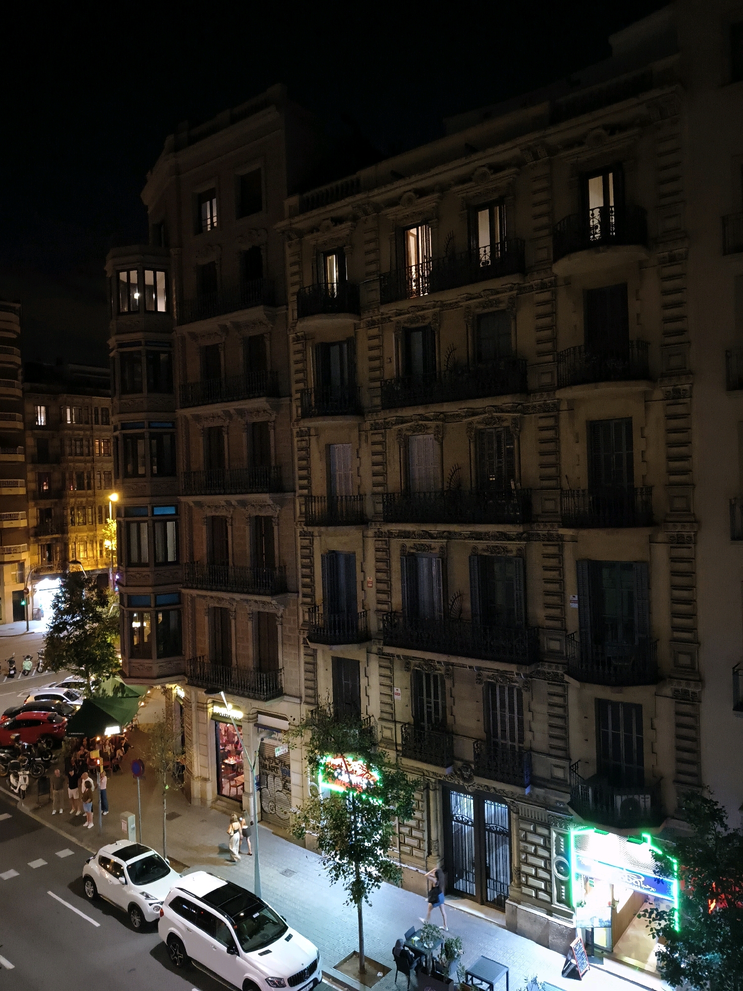 This is a night shot of a street scene in Barcelona, Spain, specifically showing a row of multi-story buildings. The architecture features ornate detailing, balconies with wrought-iron railings, and numerous windows, some illuminated from within. The street level of the buildings includes shops and restaurants, identifiable by their signs, awnings, and interior lighting. One establishment has a prominent neon green sign. Sidewalks are present, with streetlights and trees lining the street. Several cars are parked on the street, including two white SUVs, one red car, and several motorcycles or scooters. Pedestrians are visible on the sidewalks, with some standing outside a restaurant under a green awning, and others walking along the sidewalk. The overall atmosphere is that of a bustling urban environment at night.