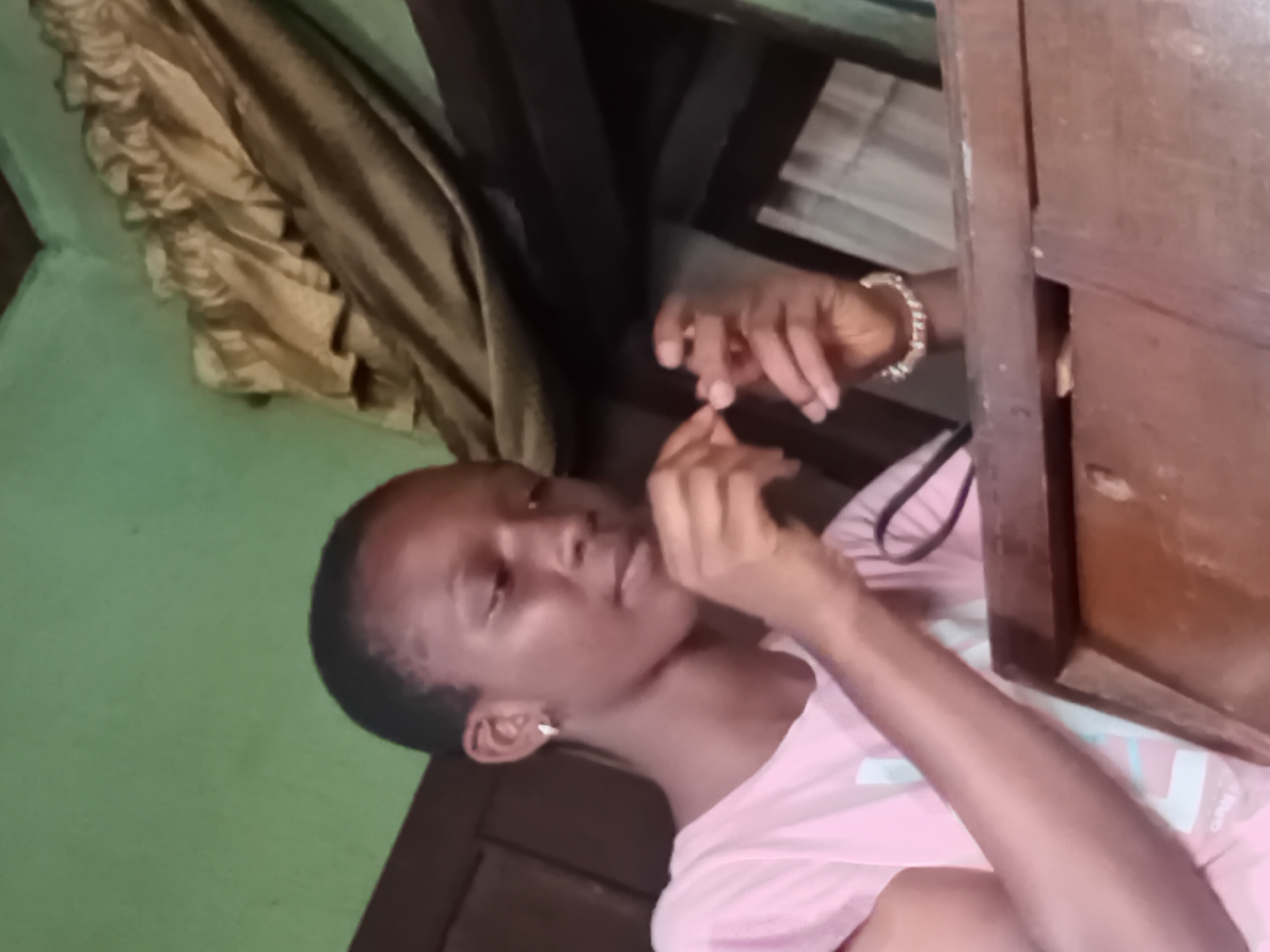 A young woman is shown in a medium shot, seated and looking down at her hands. She is wearing a pink t-shirt and a silver earring in her left ear. Her right wrist is adorned with a bracelet. Her hands are engaged in an activity, with her fingers seemingly holding a small object. The background consists of a light green wall and a section of brown curtains. To the right, a wooden surface is visible, with a drawer-like opening. The overall setting suggests an indoor environment, potentially a home, located in Nkpor, Nigeria. The lighting is dim, and the image quality is moderate.