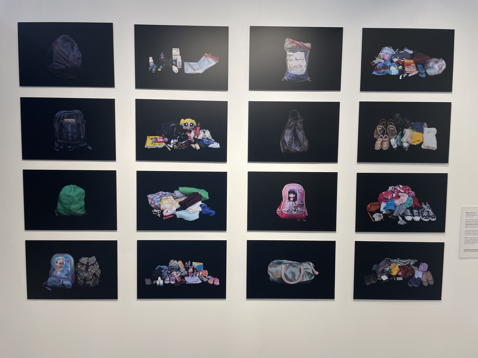 The image shows a display of sixteen framed photographs mounted on a white wall, presumably in an art gallery or museum. Each photograph depicts a different collection of personal belongings against a stark black background. The items include bags, backpacks, clothing, shoes, toys, medicine, and other miscellaneous objects. The arrangement of the photographs is in a 4x4 grid. The location is likely in Mexico City, Mexico, as the text label on the right side of the wall is in Spanish. There are no people visible in the scene, only the photographs of their possessions.