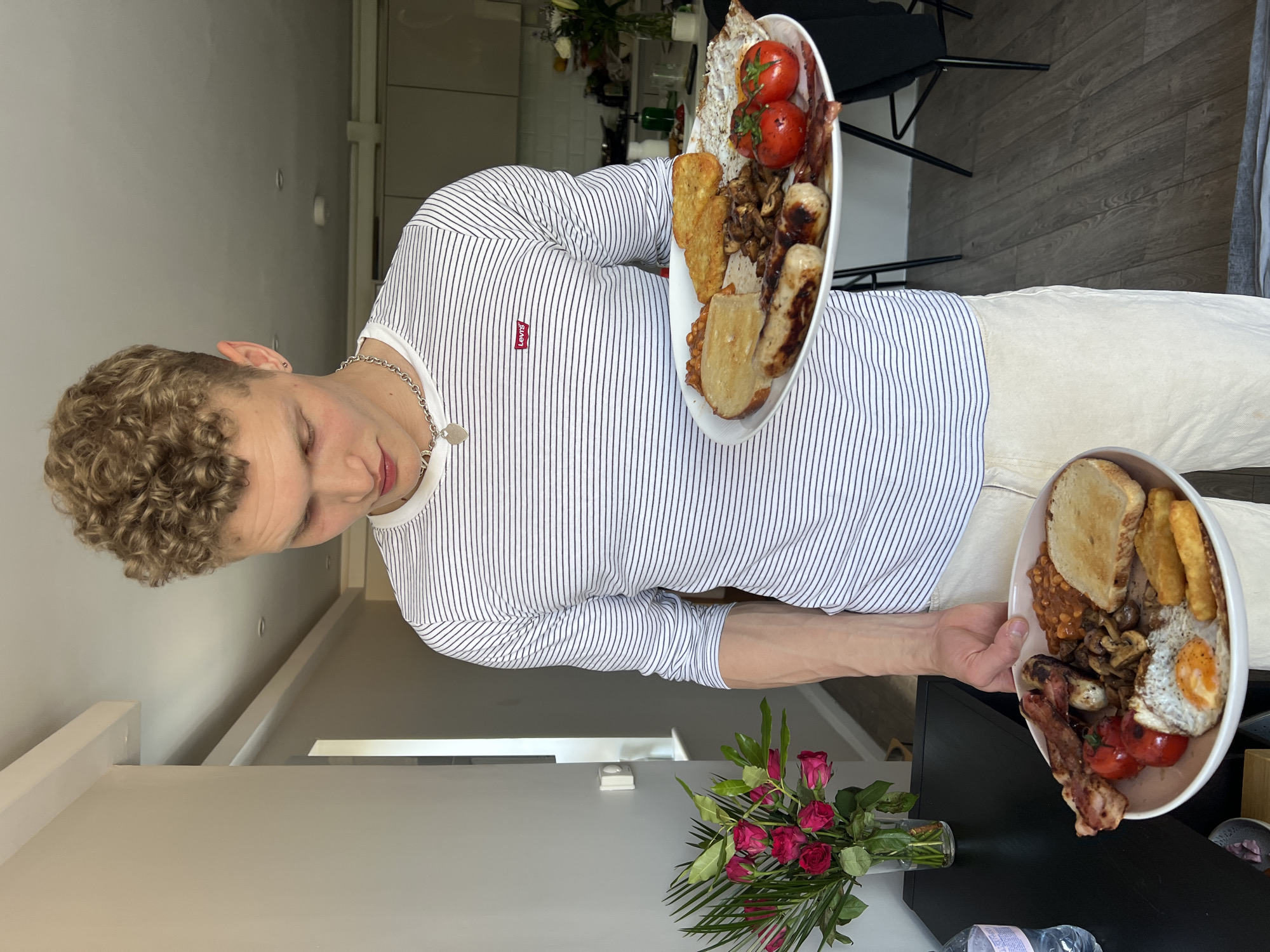 A young man with curly blond hair stands indoors, holding two plates of food. He wears a white and black striped long-sleeved shirt, light-colored pants, and a silver necklace. Each plate holds a full English breakfast including toast, fried eggs, sausages, mushrooms, bacon, baked beans, fried tomatoes, and hash browns. A vase of red roses is visible on a dark surface near the lower plate. The location appears to be a modern home, possibly in London, based on interior design and food.