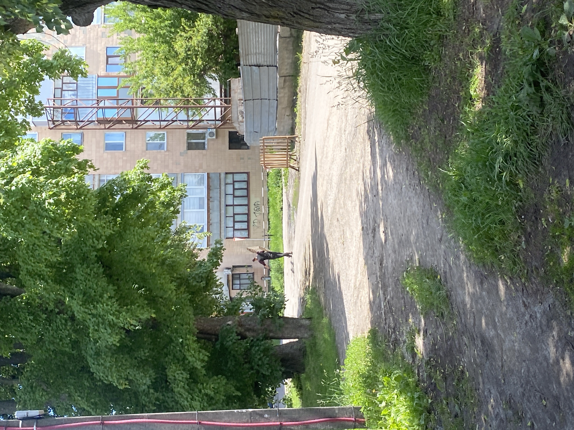This image, rotated 90 degrees counter-clockwise from its intended upright orientation, depicts an outdoor urban or suburban scene in Poltava, Ukraine, on a bright, sunny day.
A dirt and gravel path stretches from the foreground into the distance, gently curving to the right. To the left of the path stands a multi-story building with a light-colored, possibly tiled or bricked facade and numerous windows, some of which appear reflective. A prominent metal-framed structure, resembling a shared balcony or walkway, is attached to an upper level of the building, partially framed by vibrant green trees. An outdoor air conditioning unit is visible on the building's facade. Faint graffiti, possibly spelling "TRD" and "ROBA," can be seen on the lower part of the building.
Lush green trees and grass densely line both sides of the path. Large tree trunks and foliage dominate the left side of the scene, extending from the foreground upwards. In the immediate foreground, a low concrete wall or barrier with a red pipe or cable running along its top edge is visible.
In the middle distance, a person is walking along the path, away from the viewer. They are carrying a large, flat, light-colored object, possibly a wooden pallet or sheet, on their back or shoulder. Further down the path, to the right of the building, there are stacks of corrugated sheets, likely building materials, and a small wooden barrier or gate.
The scene is illuminated by strong sunlight, casting shadows on the path and grass, indicating clear weather. The overall impression is one of everyday activity in a lived-in environment combining natural elements with urban architecture.