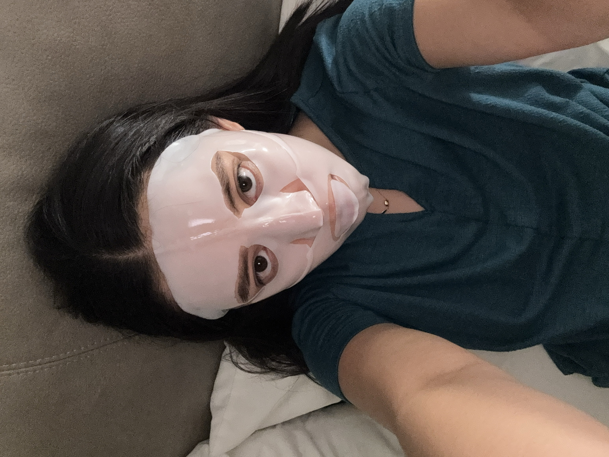 A woman with long, dark hair is lying on a gray couch with white pillows, wearing a turquoise V-neck shirt and a white facial sheet mask. The scene appears to be indoors, possibly in a home setting. There are no discernible landmarks or details to indicate the specific location within Puebla, Mexico.