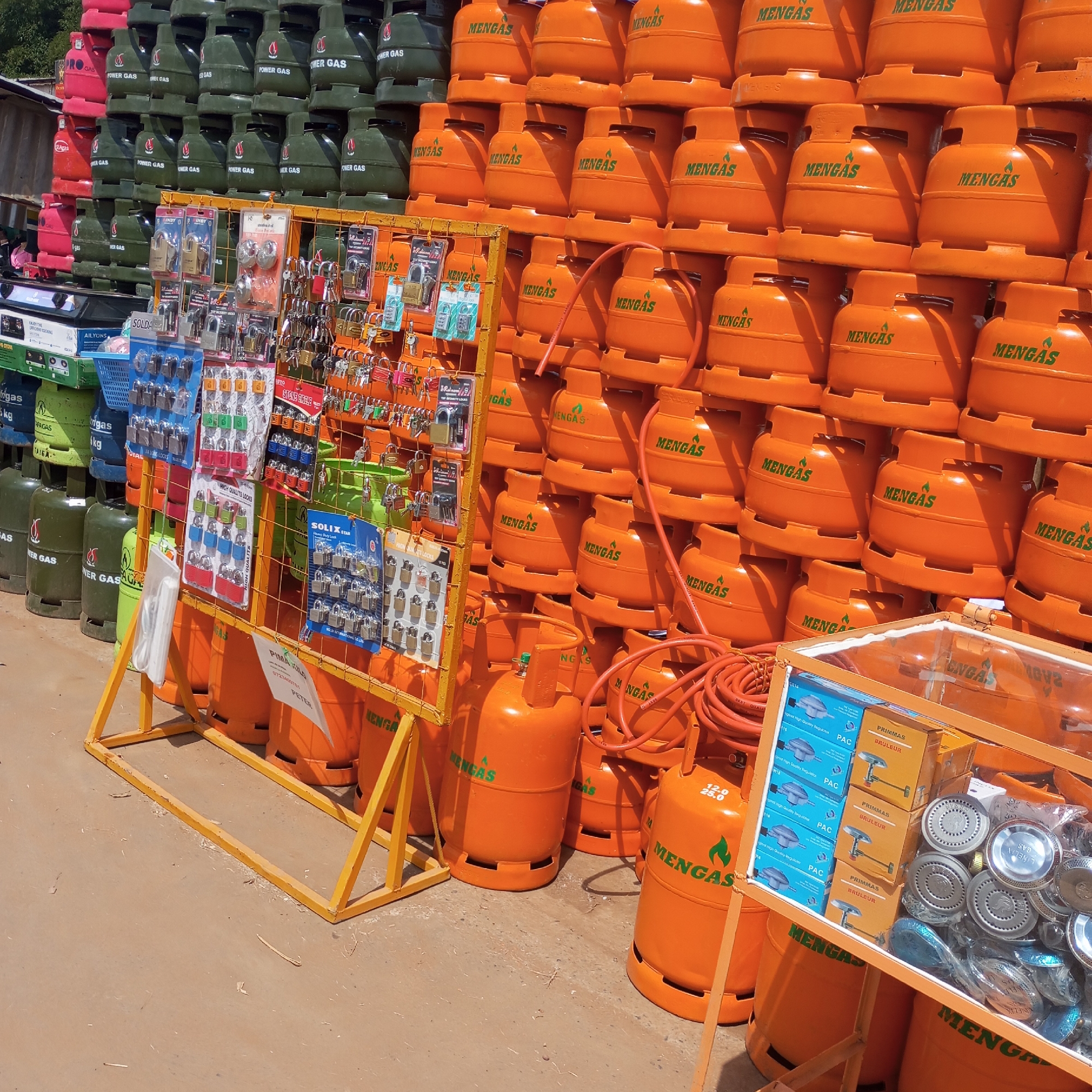 The image shows a roadside shop selling gas cylinders in Sheywe ward, Kenya. Numerous gas cylinders, stacked in rows, dominate the scene. These cylinders are branded with "MENGAS" and "POWER GAS". Some are green, while others are orange. A yellow metal rack displays a variety of padlocks for sale. Accessories such as gas regulators and burners are presented in a glass display case next to the cylinders. The ground is dusty. There are no people visible in the immediate frame.