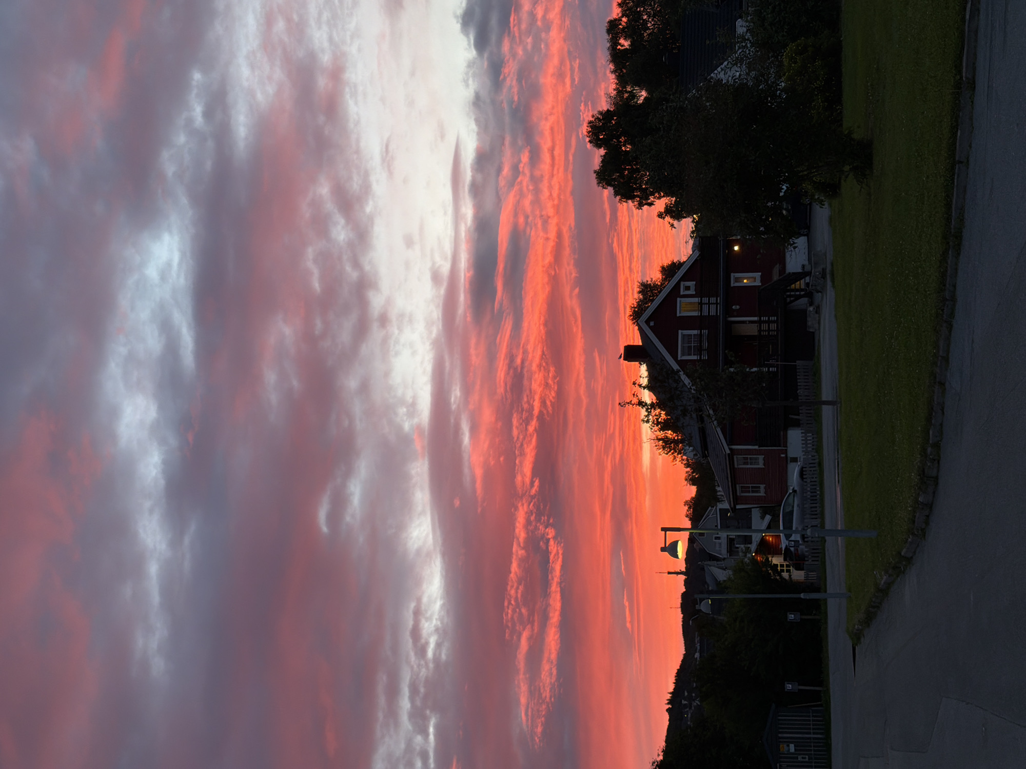 The image depicts a sunset over a residential area in Harstad, Norway. The sky is filled with vibrant red and pink clouds, tinged with grey and white streaks, indicative of the twilight hours. In the foreground, a paved road curves to the left. Adjacent to the road is a grassy area with a stone border. Several streetlights are visible, casting a faint glow. A red house with white trim and multiple windows is prominently featured, with a dark-colored roof and a porch. A silver car is parked in the driveway of the house. Behind the house, dark green trees create a natural backdrop. In the distance, hills can be seen, and other buildings are sparsely scattered across the landscape. The overall scene suggests a peaceful evening in a suburban or semi-rural setting.