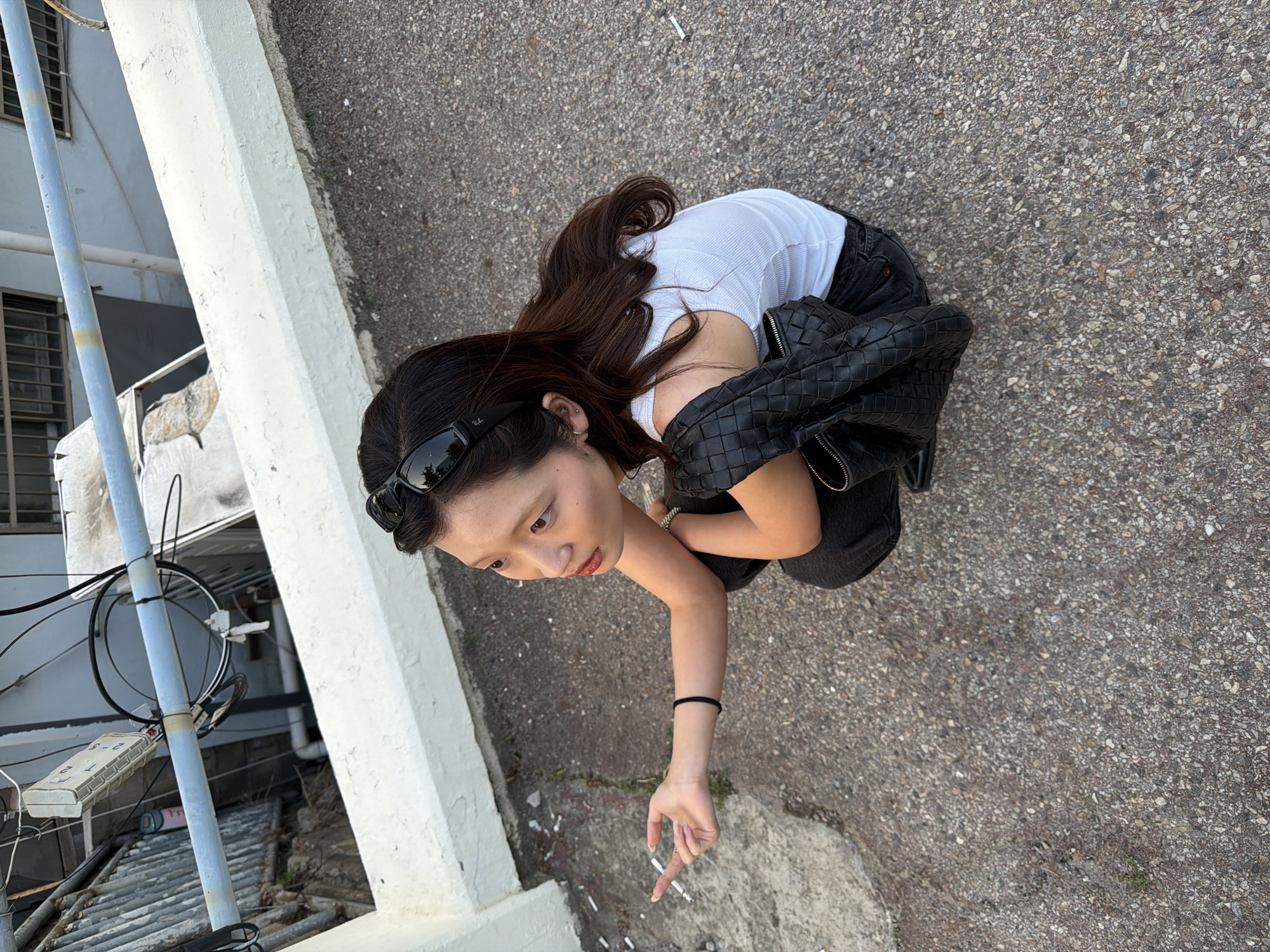 A woman in Seoul, South Korea, is standing on a concrete surface littered with cigarette butts. She is wearing a white tank top, black jeans, and sunglasses perched on her head. A black leather bag hangs off one shoulder. Her arm is extended, and her fingers are near a cigarette butt. To the left is a building with exposed pipes and a white concrete ledge. The surface is likely a street or alleyway. The time of day appears to be daylight.