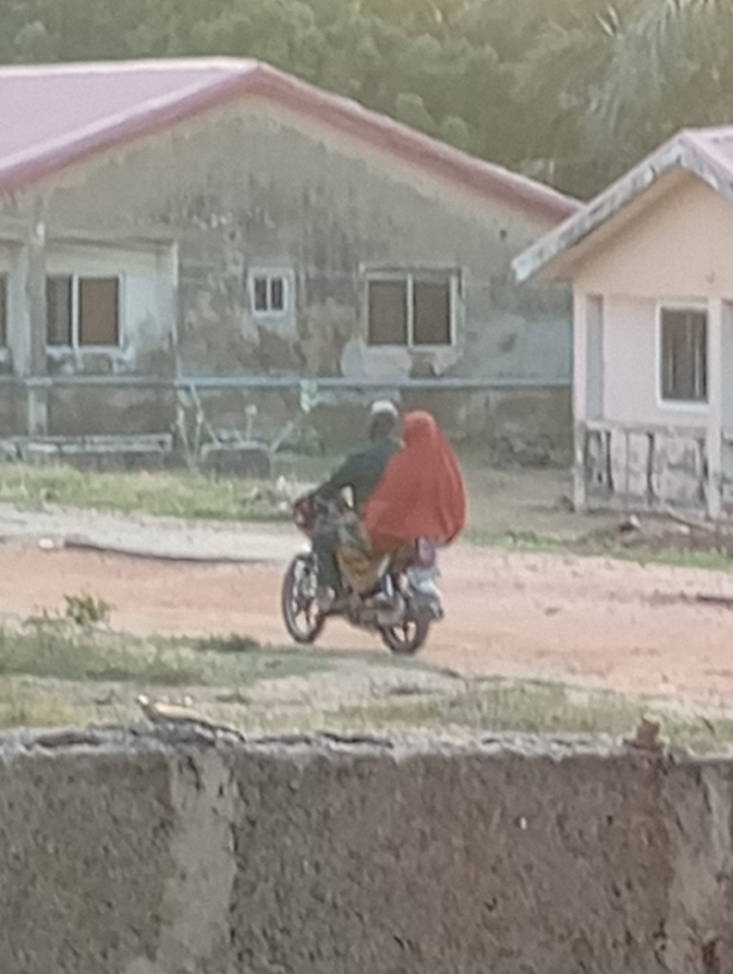 Two people are riding a motorcycle on a dirt road in front of two buildings. The person in front is wearing a dark shirt and a light-colored cap. The person in the back is wearing a red hijab. The buildings appear to be stucco with tiled roofs. The building on the left has several windows, and the building on the right also has windows and a porch. There is some greenery and trees in the background. In the foreground, there is a stone wall.