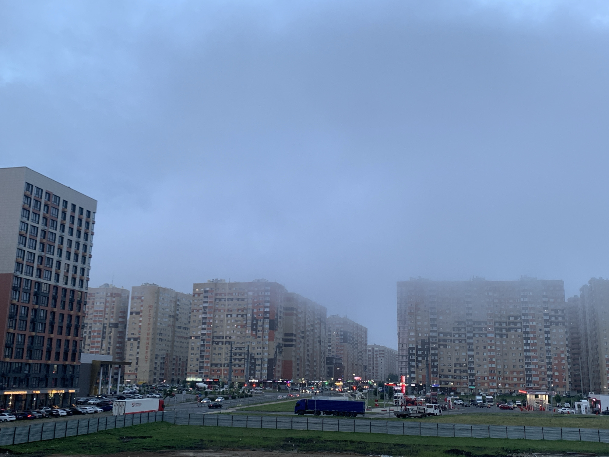 The image depicts a cloudy, overcast sky over a city street scene. The scene features several tall residential buildings with visible windows and balconies, some of which are partially obscured by fog or low clouds. A multi-lane road runs through the foreground, with various vehicles, including cars, trucks, and possibly construction equipment, present. There are streetlights and some signs of commercial activity, such as what appears to be a gas station or a series of shops. The overall atmosphere suggests an urban environment during twilight or a heavily overcast day. The buildings are a mix of light-colored facades with some darker accents. The image is captured from a slightly elevated viewpoint looking down towards the street and buildings.
