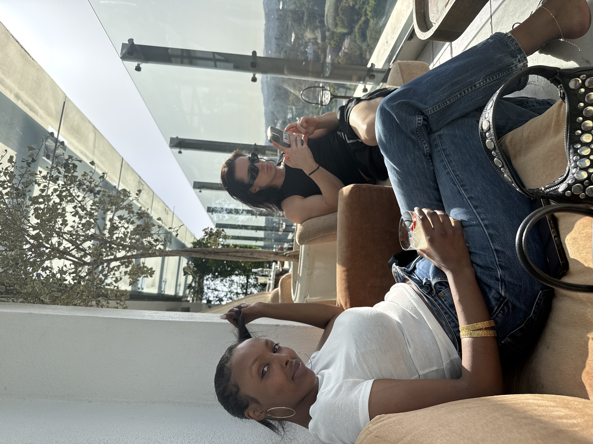 Two women are relaxing outdoors in what appears to be a rooftop bar or lounge in West Hollywood, California. The younger woman is lying back in an upholstered chair, holding her hair up with one hand. She is wearing a white t-shirt, blue denim jacket, and blue jeans, with a pair of gold bracelets on her wrist. She is holding a cocktail in her other hand, and is looking directly at the camera with a slight smile. The second woman is sitting upright in a similar chair behind the first woman, looking at her cell phone. She has long, dark hair and is wearing dark sunglasses and a dark, sleeveless top. A glass safety barrier is visible in the background, overlooking a cityscape with hills and vegetation.