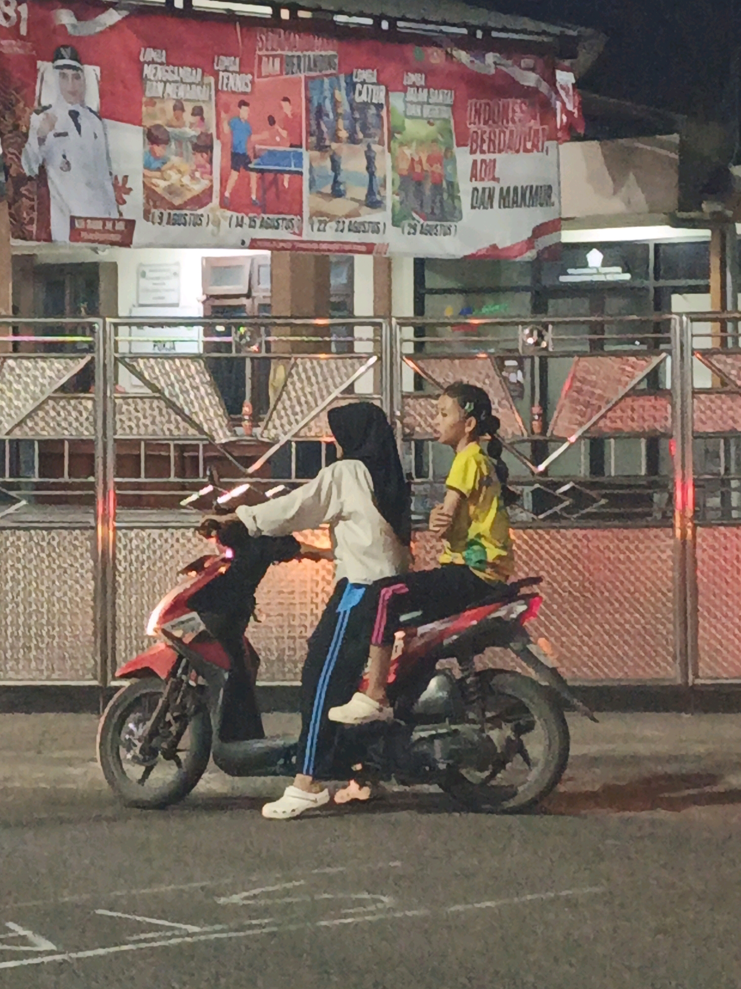 In Ketegan, Indonesia, two young females are captured in a side-profile view, seated on a red and black motorcycle. The individual in front, wearing a yellow t-shirt and black pants, has her hair tied in a braid and is looking to her left. The person behind her, wearing a white long-sleeved shirt and black pants with blue stripes, is also looking to her left. Both are wearing casual footwear. The motorcycle has a red front fairing and a black body. The background features a metal fence with an intricate geometric pattern and a banner displaying multiple images and text in Indonesian. The banner shows a man in a white uniform, images related to sports like table tennis, and text that appears to be related to events or a celebration. The lighting suggests it is evening or night, with some artificial lights reflecting on the scene.
