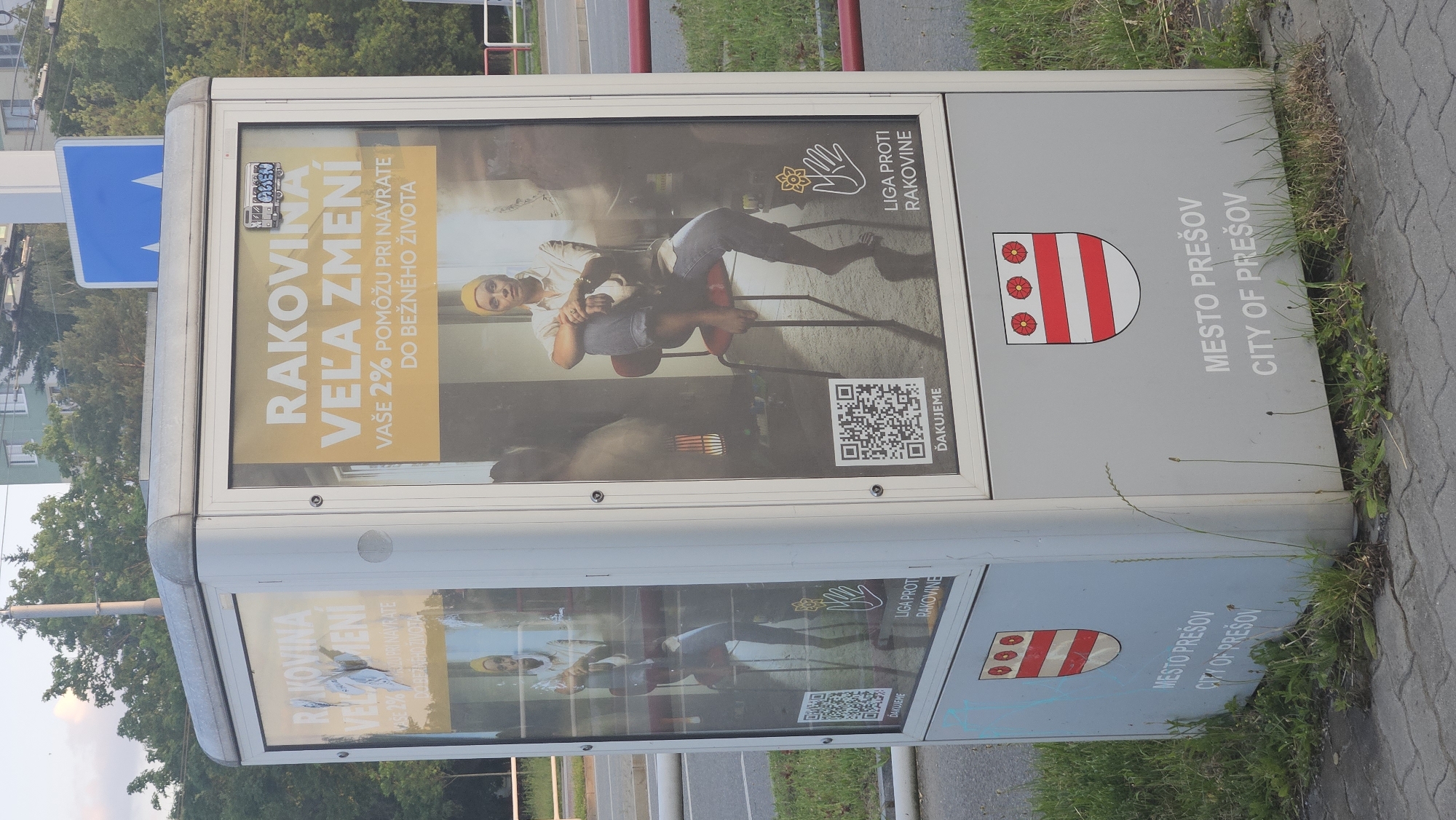 The image shows a public advertising kiosk in the city of Prešov, Slovakia. The kiosk is a rectangular structure with glass panels displaying advertisements. On the front panel, there is a large yellow advertisement with the text "RAKOVINA VEĽA ZMENÍ" (Cancer Changes a Lot) and "VAŠE 2% POMÔŽU PRI NÁVRATE DO BEŽNÉHO ŽIVOTA" (Your 2% will help you return to normal life). Below this text, there is an image of a woman wearing a yellow headscarf and jeans, sitting on a chair. To the left and right of the woman, there are QR codes and the logo of "LIGA PROTI RAKOVINE" (League Against Cancer). The kiosk also features the coat of arms of Prešov, which is a shield with red and white stripes and three red roses on top. The text "MESTO PREŠOV" and "CITY OF PREŠOV" are visible on the lower part of the kiosk. The kiosk is situated on a sidewalk, with grass and weeds growing around its base. In the background, there are trees, buildings, and a road.