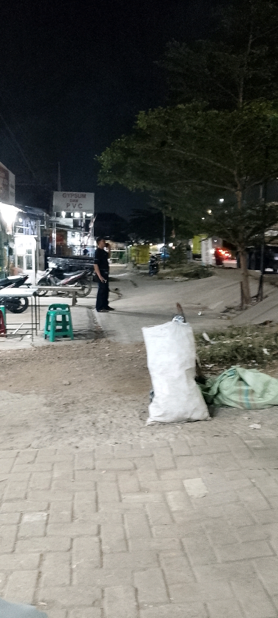 The image captures an outdoor scene at night in Cileungsi, Indonesia, likely a commercial area given the presence of signage for "GYPSUM DAN PVC." A lone individual, appearing to be a security guard or worker, stands near a row of shops, looking towards the left of the frame and holding something to their mouth. In the foreground, a green plastic stool is positioned on a paved walkway. Adjacent to the walkway, on a dirt and grass area, are two large bags: one white and rectangular, and another lighter colored bag to its right. A tree with dense foliage is visible in the background, with its branches extending into the dark night sky. The environment suggests a street or alleyway adjacent to businesses, illuminated by artificial lighting, with motorcycles and other vehicles partially visible in the distance.