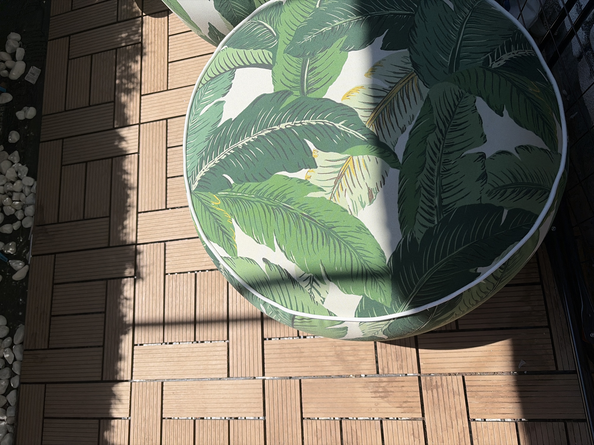A circular cushion with a tropical leaf pattern is placed on a wooden deck. The deck is made of rectangular planks arranged in a staggered pattern. Sunlight casts shadows across the deck and onto the cushion. Small white stones are visible in the upper left corner of the image. The scene appears to be an outdoor setting.
