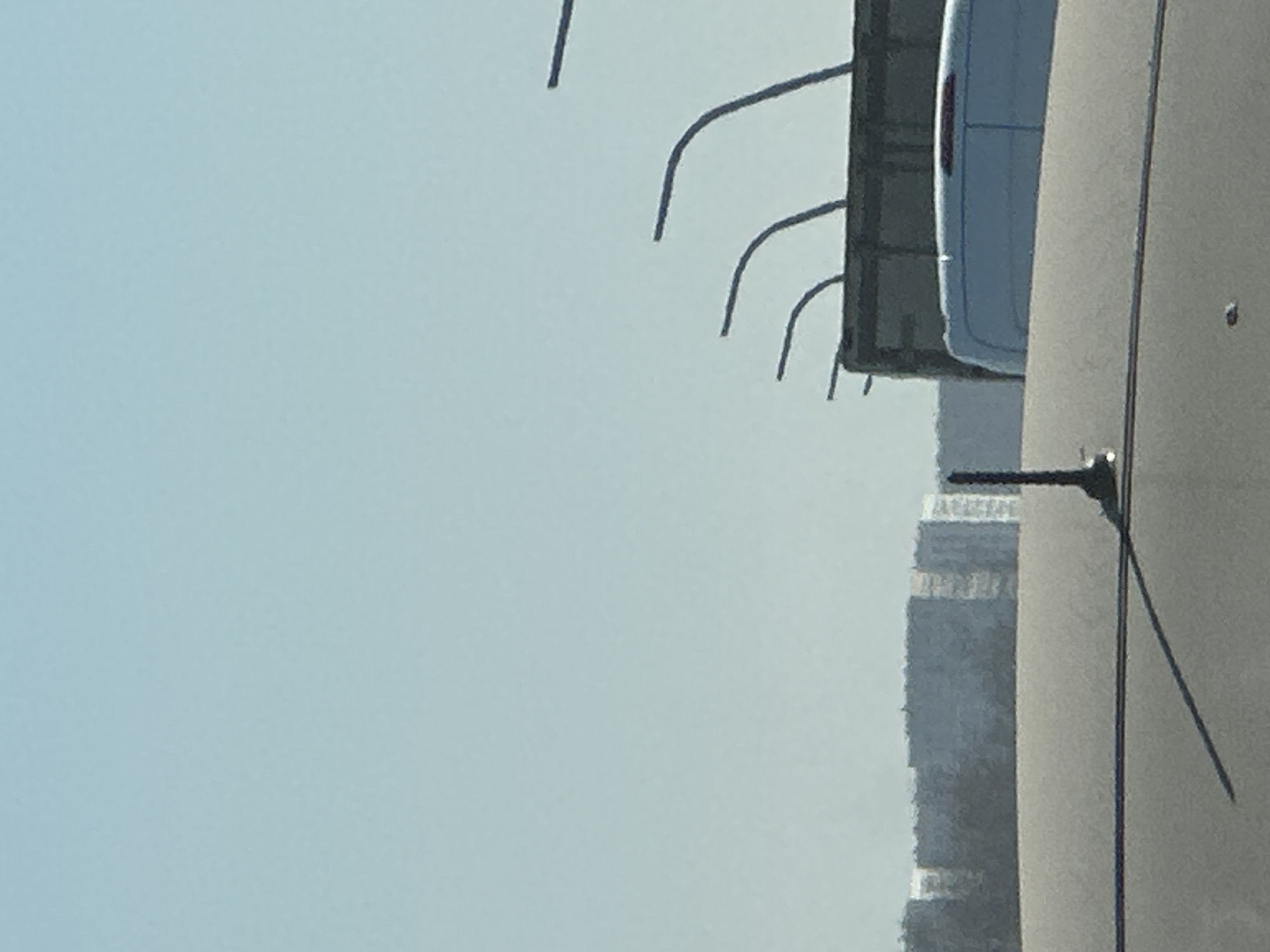 The image depicts a scene in what appears to be an urban environment. The sky is clear and light blue. A portion of a white vehicle with a red taillight is visible on the upper right side of the frame, situated next to a gray concrete surface. Overlapping the vehicle are a series of black, curved rods. Below the road, a building with multiple layers is on the right of the image. A tall, black pole casts a shadow across the concrete surface. There are no people visible in the image. The location cannot be determined from the image.