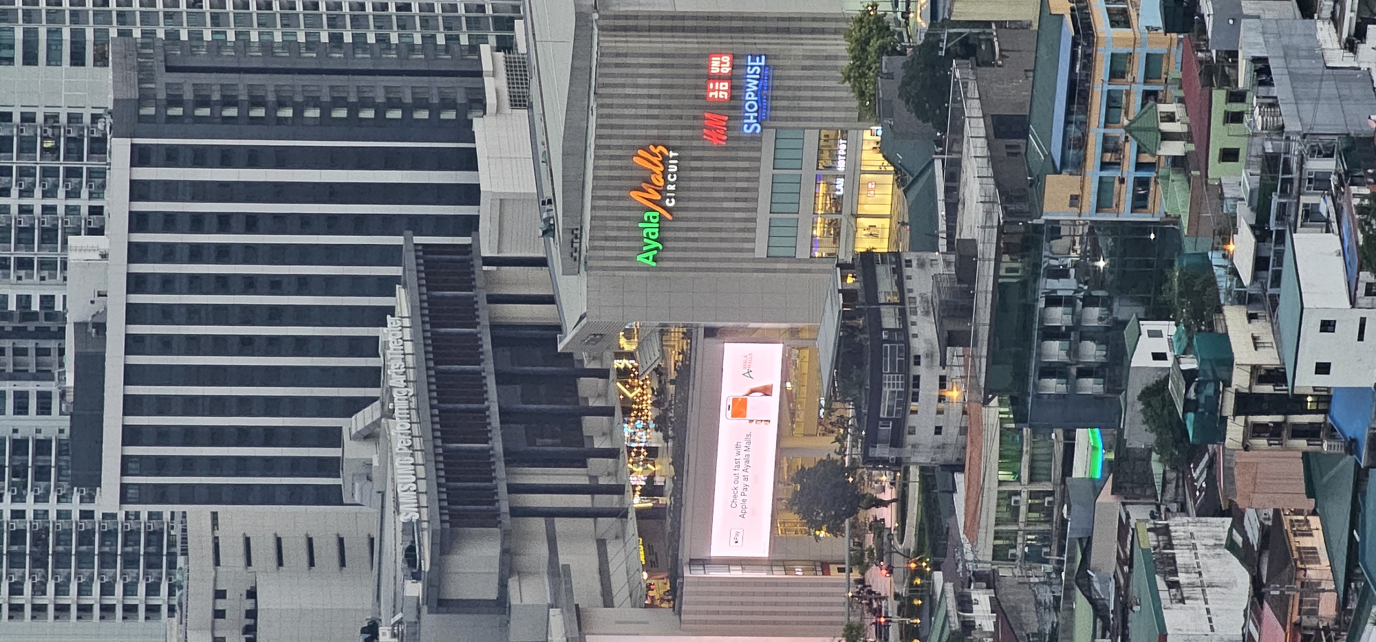 This is an aerial view of the Ayala Center area in Makati City, Philippines, likely captured during the daytime with overcast or dusk conditions. The scene is dominated by a dense collection of modern high-rise buildings and lower-rise structures.
Prominently featured is the Ayala Malls Circuit building, identifiable by its green "Ayala Malls" logo and vertical striped facade. Adjacent to it are signs for H&M, UNIQLO, and SHOPWISE. A large digital billboard displays an advertisement for Apple Pay at Ayala Malls. Further left, the Samsung Performing Arts Theater is visible.
In the foreground and middle ground, a cluster of older, lower-rise buildings with multi-colored facades and corrugated metal roofs are visible, indicative of a more densely populated urban area. Some buildings have visible air conditioning units, and a few appear to be under construction or renovation, with green netting covering parts of them.
The overall impression is of a bustling, multi-layered urban environment characteristic of a major business and commercial district. There are no visible people in the image, and the focus is on the architecture and urban landscape.