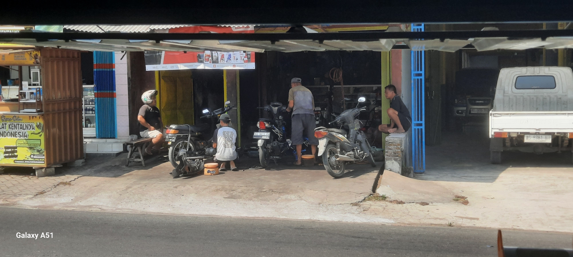 This is a medium-angle, eye-level shot taken from an elevated position looking down into an open-air workshop in Selopuro, Indonesia. The scene depicts a motorcycle repair shop with several individuals and vehicles present.
In the foreground, a concrete road is visible, with the bottom edge of the image framing the scene. To the left, a brightly colored stand with yellow and green signage is present, featuring text in Indonesian, including "Croco Celum" and "LAT KENTALNYA INDONESIA." This stand appears to be a food or beverage vendor.
The workshop itself is partially covered by a corrugated metal roof supported by dark beams. The ground inside the workshop is paved with concrete. In the center of the frame, two men are working on a motorcycle. One man, wearing a white t-shirt with a complex graphic print and a green cap, is crouched down, appearing to be actively engaged in repairs. Another man, wearing a blue and grey shirt and grey shorts, stands behind the motorcycle, also seemingly involved in the repair process.
To the left of the main repair area, a man wearing a black t-shirt, white shorts, and a white motorcycle helmet is seated on a wooden bench. He is looking towards the right side of the frame.
On the right side of the workshop, a young man is squatting on a low brick wall, looking to his left. He is wearing a dark t-shirt and dark shorts.
Several motorcycles are parked in the workshop. The one being repaired in the center is black and has a license plate that reads "AG 3673 PH." Another motorcycle, primarily white with black accents, is parked to its right. A third motorcycle, mostly black, is partially visible behind the white one.
Further into the background, to the right, the rear of a white pickup truck is visible, parked in what appears to be a covered area. This truck has a license plate that reads "AG 9781 FJ." In the darker background of the workshop, other vehicles, including what appears to be an SUV with a license plate "AG 1855 PF," are parked.
The environment is characterized by bright daylight, casting distinct shadows. The overall impression is of a busy, functional roadside workshop.
