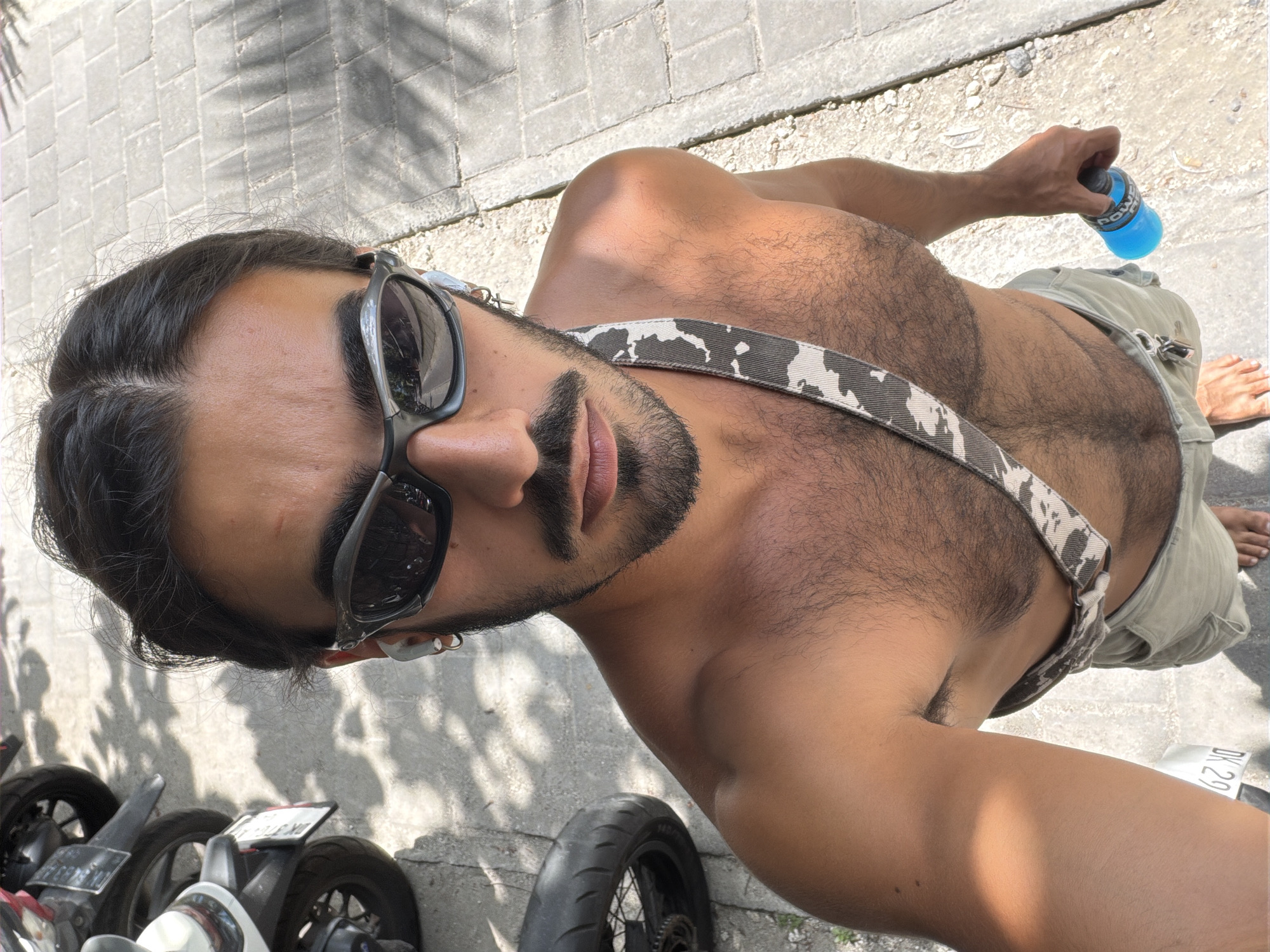 A shirtless person wearing sunglasses and khaki shorts stands outdoors, holding a blue bottle. They have a patterned strap across their chest and are wearing white earbuds. Behind them, the tires and license plates of several motorcycles are visible. The ground is paved with stone. The scene appears to be in a city, potentially North Kuta, Indonesia, given the presence of motorcycles and tropical vegetation creating dappled shade.
