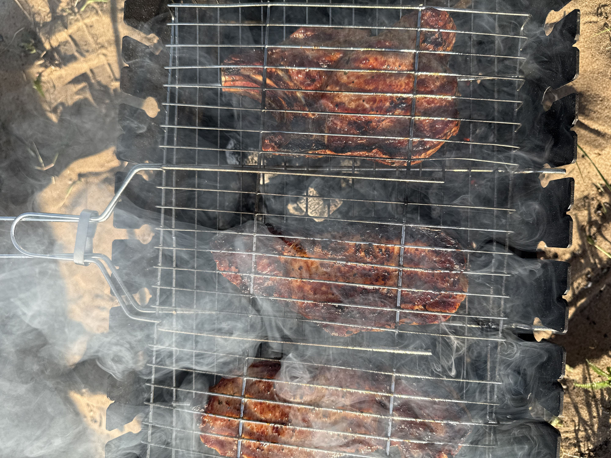 The image shows three pieces of meat cooking on a metal grill. The grill is emitting smoke. The background shows sand and some vegetation. The location is Chaikovsky, Russia.