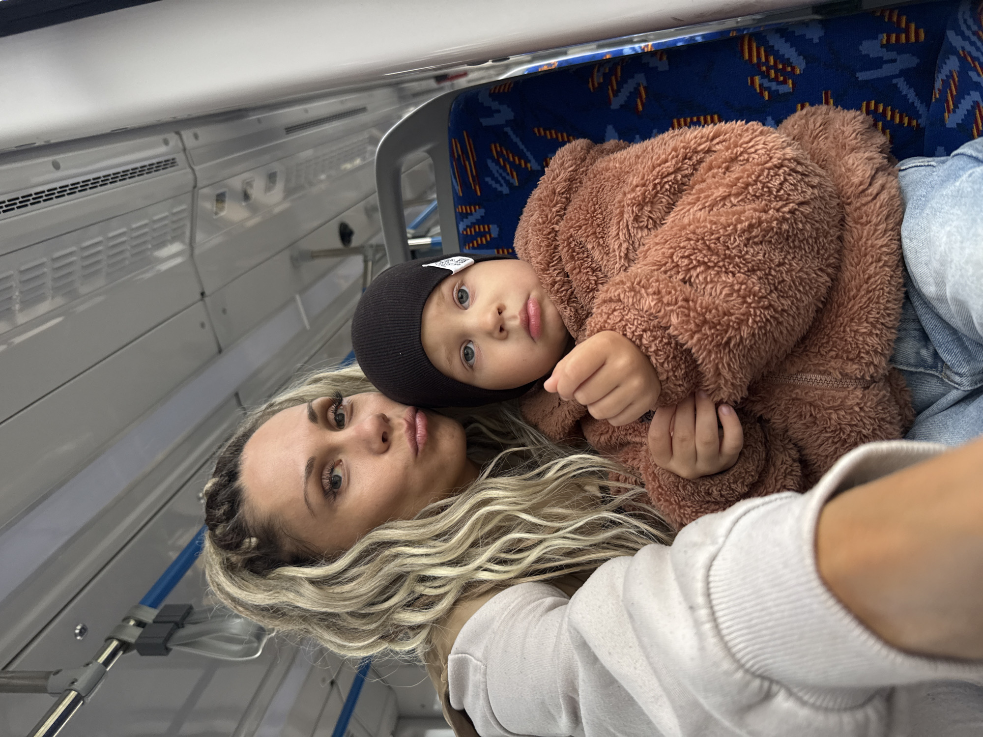 A woman and a child are sitting on a bus seat in Rusanovo, Russia. The woman is on the left, and the child is on the right. The woman has blonde hair and is wearing a light-colored sweatshirt. She is making a duck face. The child is wearing a black beanie and a brown fuzzy jacket. The child is looking at the camera with a neutral expression. The bus seats are blue with a pattern of red and yellow. The interior of the bus is visible in the background, with overhead compartments and handles.