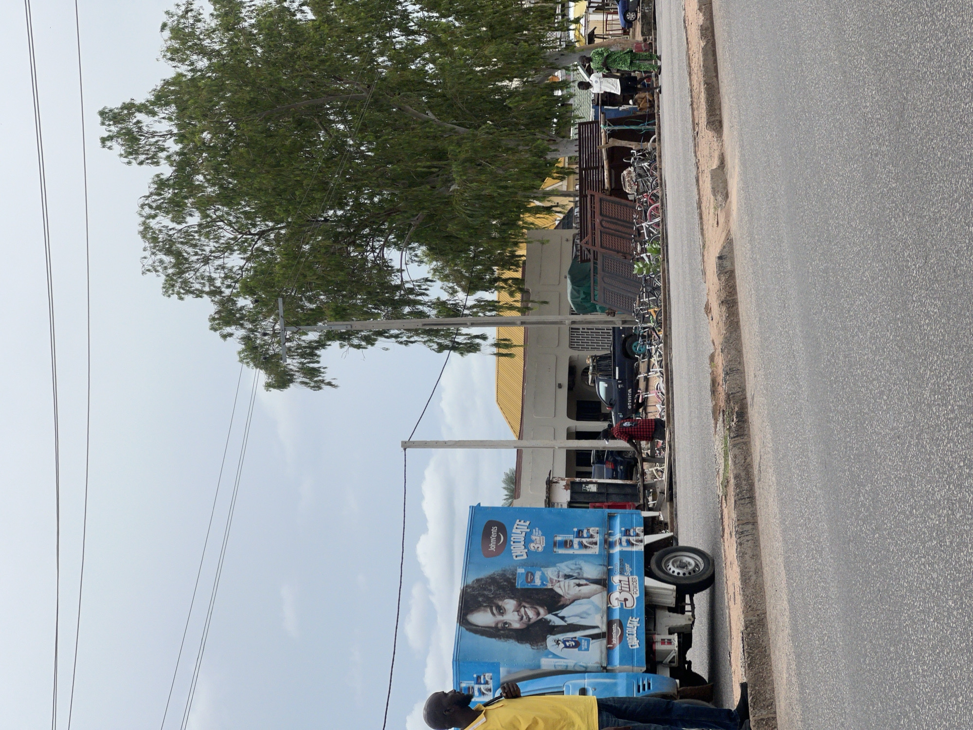 In Jalingo, Nigeria, on a street with a paved road and a raised sidewalk, a blue truck with an advertisement for "John Vents Chocolate 3in1 Powder" featuring a smiling woman is parked. To the left of the truck, a man in a yellow shirt stands facing the truck. Behind the truck, a collection of bicycles are displayed for sale. Several people are present: two men in the foreground near the bicycles, one in a green patterned garment and the other in a white shirt and black shorts, and another person wearing a red and black checkered shirt to the right of the bicycles. In the background, buildings with yellow roofs and a large tree with dense green foliage dominate the scene. Overhead, power lines crisscross the overcast sky.