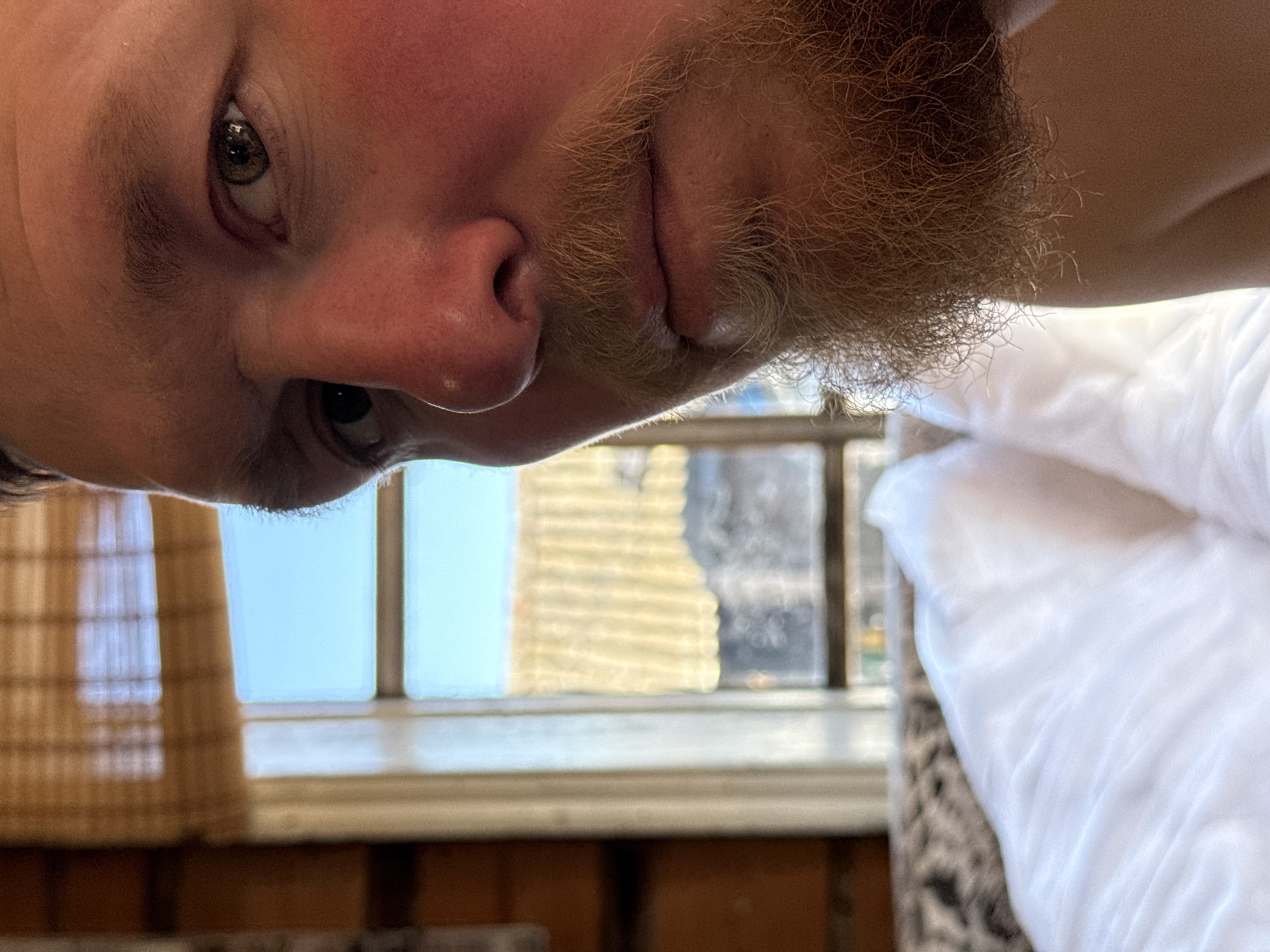 A selfie shot from a high angle shows a man's face, with his right eye visible and gazing downward. He has a fair complexion and a ginger beard. The background features a window with partially drawn blinds, revealing an out-of-focus building exterior. A white blanket and patterned fabric are also in the background. This location cannot be determined as being in Rosstani, Russia, based on visual information.