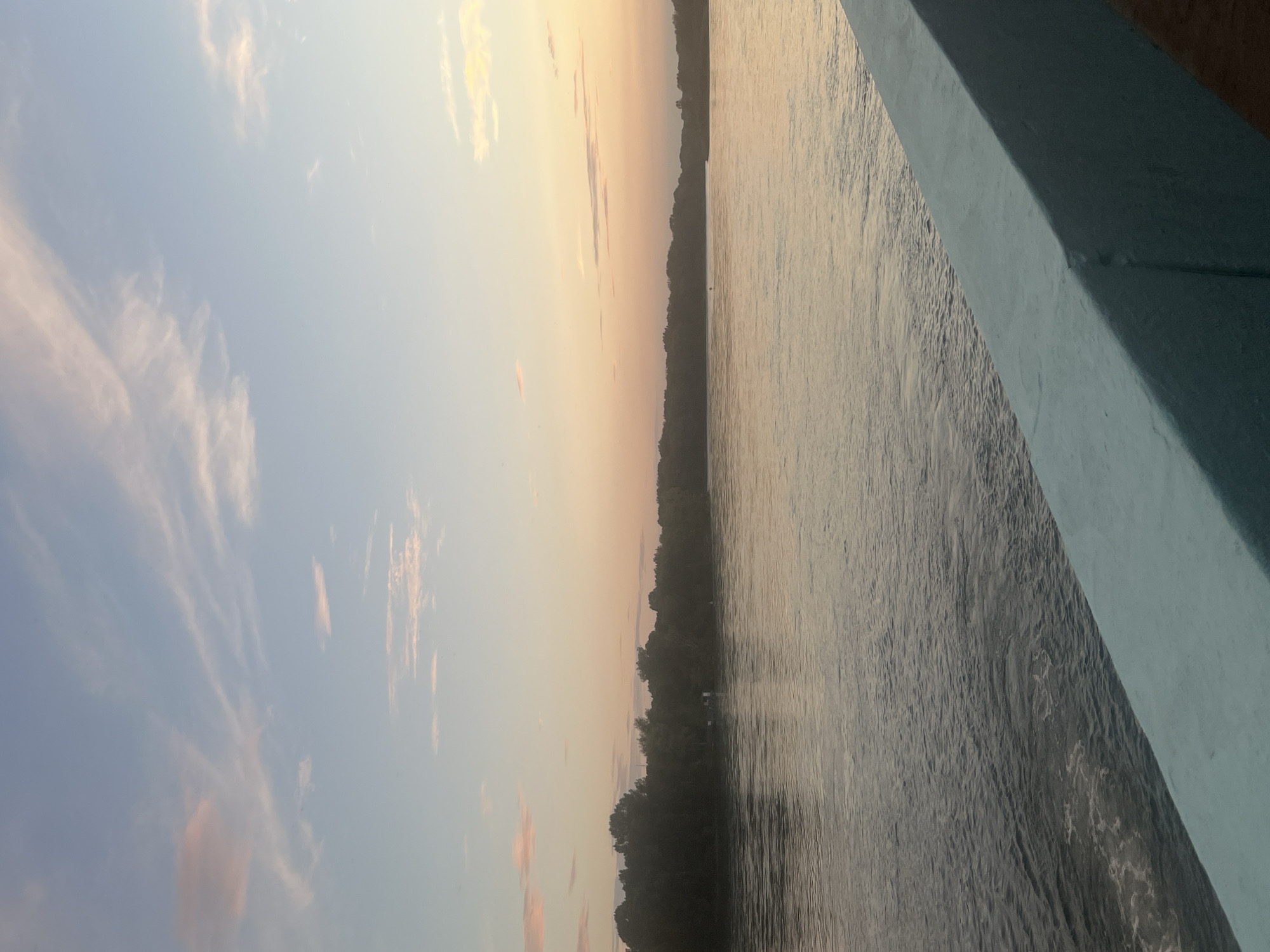 A scenic view of a wide river at sunset, as seen from a bridge. The water is calm with gentle ripples, reflecting the soft colors of the sky. A dense line of trees forms the distant shoreline. The sky is a gradient of pale blue at the top, transitioning to warm hues of pink and orange near the horizon, with scattered wispy clouds. The foreground features the light blue railing of the bridge. There are no people visible in the image. The location is not specified in the image or its context.