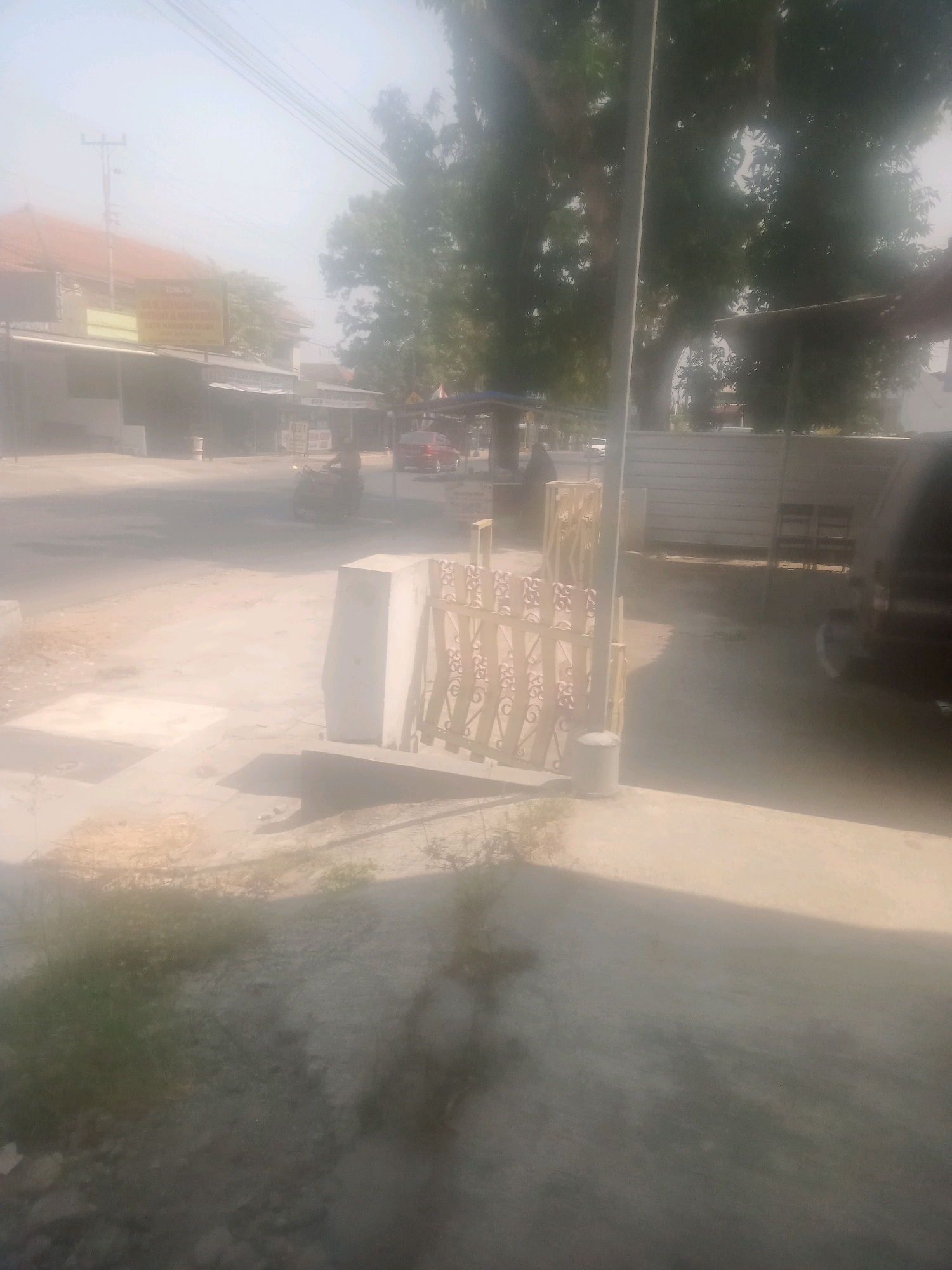 The image captures a street scene in Lebaksiu, Indonesia, possibly on a hazy or dusty day. In the foreground, there is a concrete structure with a decorative metal gate. To the left, a patch of dry grass and dirt is visible. Further back, a dirt road curves into the distance. On the left side of the road, buildings with awnings and signage are present. A red car is seen driving on the road, and a person on a motorcycle is also visible. In the background, large trees with dense foliage line the street. The sky is light and hazy, suggesting poor visibility.