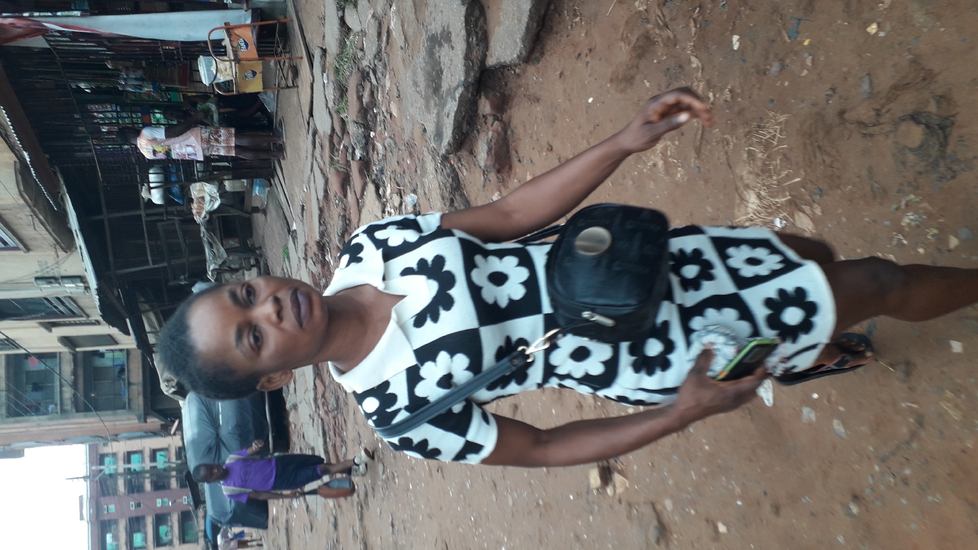 A woman with short black hair stands on a dirt and concrete street in Onitsha, Nigeria. She wears a black and white flower-patterned dress and a black bag across her chest. She is holding a cell phone. In the background, there are buildings, a makeshift market stall with people, and other pedestrians. The ground is a mix of dirt and broken concrete.