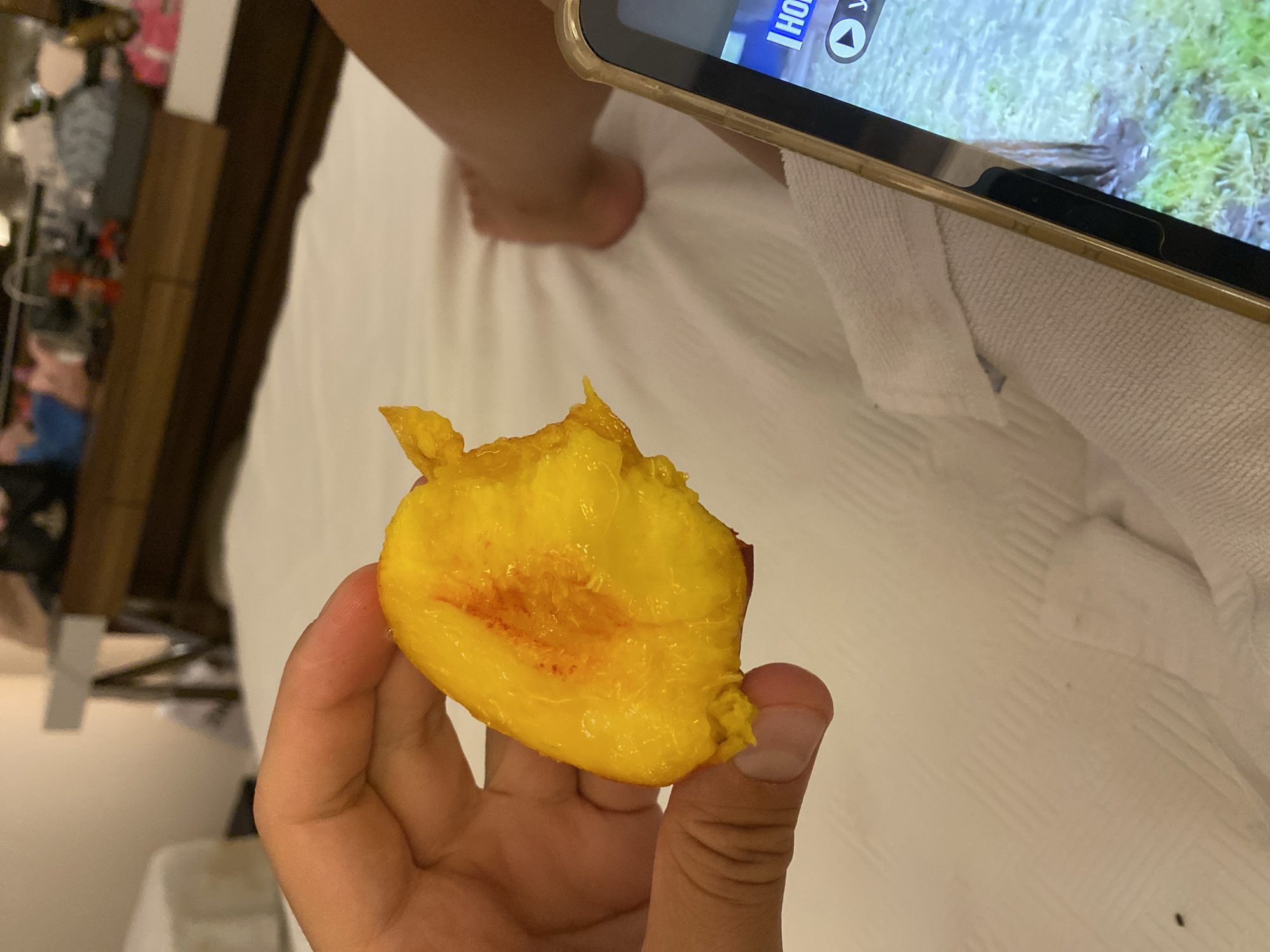 A person's hand holds a bite of a peeled peach over a white bed. A smartphone is partially visible in the lower right corner, displaying a video. The background shows a dresser with items on it and a portion of another bed. The scene appears to be indoors, likely in a hotel room. The location is not specified in the image or its metadata.