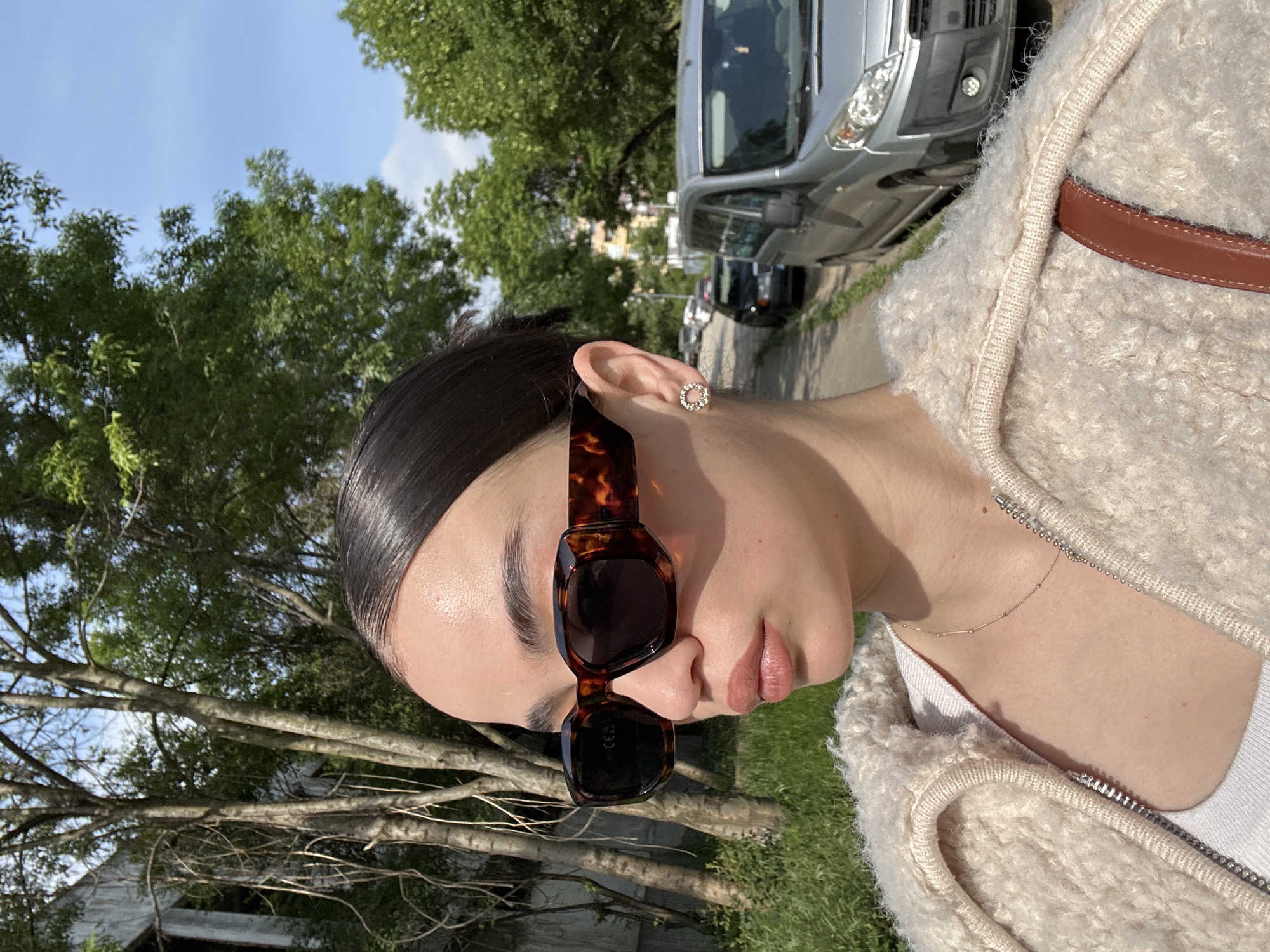 A woman is wearing sunglasses, a tan fluffy jacket with a brown leather strap, and a gold necklace. She has her hair slicked back and is wearing round, bejeweled earrings. The background shows trees, grass, a street, and cars. The location is possibly in Sofia, Bulgaria.