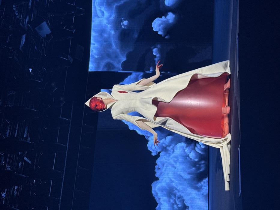 A figure stands on a stage against a backdrop of projected clouds. Dressed in a long white coat with a hood, they wear a red mask. Underneath the coat, a red garment extends from the waist, creating a wide, unusual shape. Long, claw-like fingernails are visible on their hands. The lighting is dramatic, with the figure standing out against the blue background. Stage lights are visible in the dark areas.