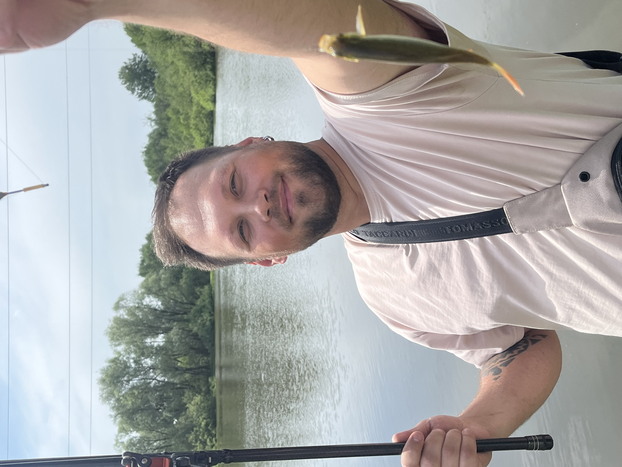 A man is holding up a small fish in an outdoor setting, likely a lake or river. He is wearing a light pink t-shirt and a shoulder bag with the strap labeled "TACCARDI TOMASS". He has a beard, short dark hair, and an earring. He is holding a black fishing rod in his other hand. The background consists of water, trees, and a partly cloudy sky. The specific location, Фоки, Russia, is not discernible from the image.
