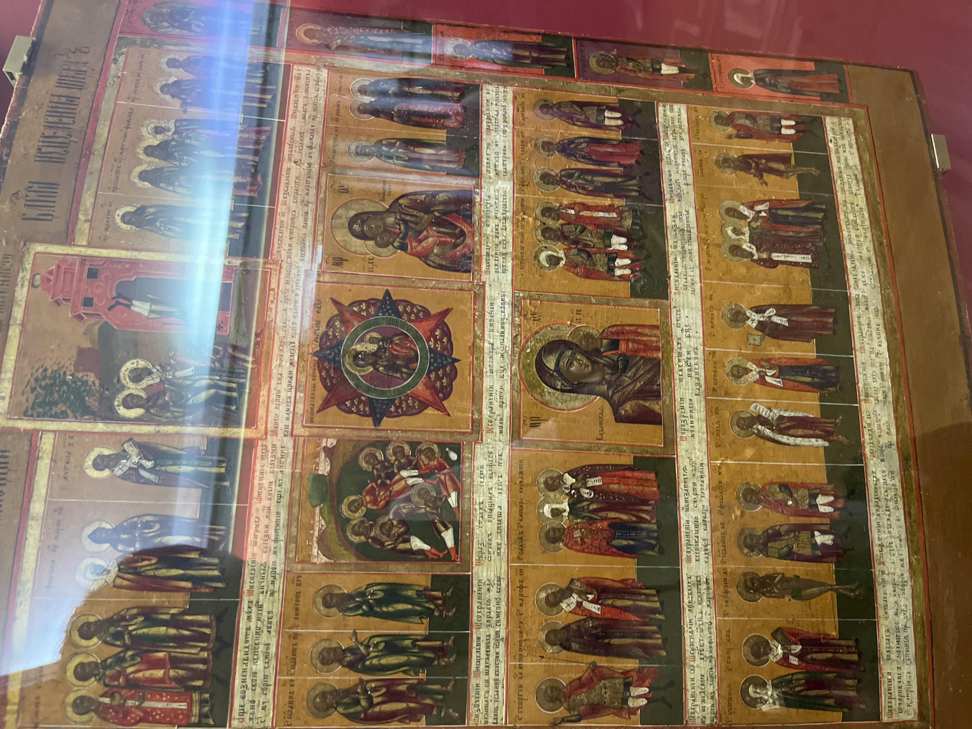 This is a photograph of a large, ornate, framed religious icon. The icon is rectangular and appears to be made of wood, with multiple painted panels depicting various saints and biblical scenes. The surface of the icon is weathered and has some visible cracks and imperfections. There are handwritten inscriptions in Cyrillic script above and below many of the painted panels. The icon is displayed in what appears to be a museum or gallery setting, with a red background visible behind and around it. The lighting suggests the photo was taken indoors, possibly with some reflections on the protective glass. Based on the style of the icon and the inscriptions, it is likely of Eastern Orthodox origin. Without further information or context, it is impossible to determine its specific location within the city of Brest, Belarus.