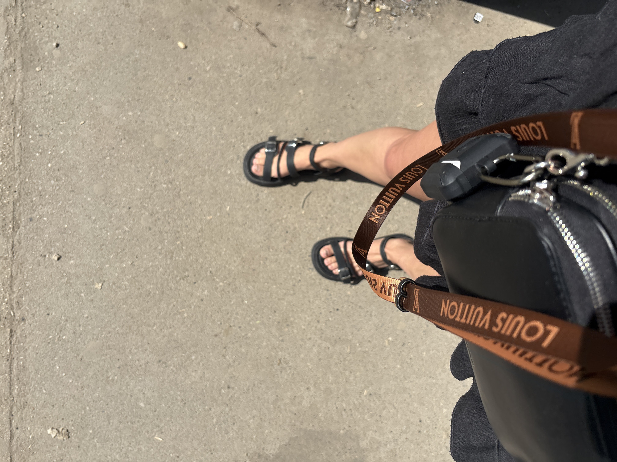 A high-angle shot depicts a person standing on a concrete surface, likely a sidewalk, based on the texture and color. The person wears black sandals with multiple straps and a black garment, possibly a dress or shorts. A black bag with a brown Louis Vuitton-branded strap is slung over their shoulder, partially obscuring their body. The setting is an urban environment, indicated by the concrete and a few pieces of debris visible on the sidewalk. The image was taken in Perm, Russia.
