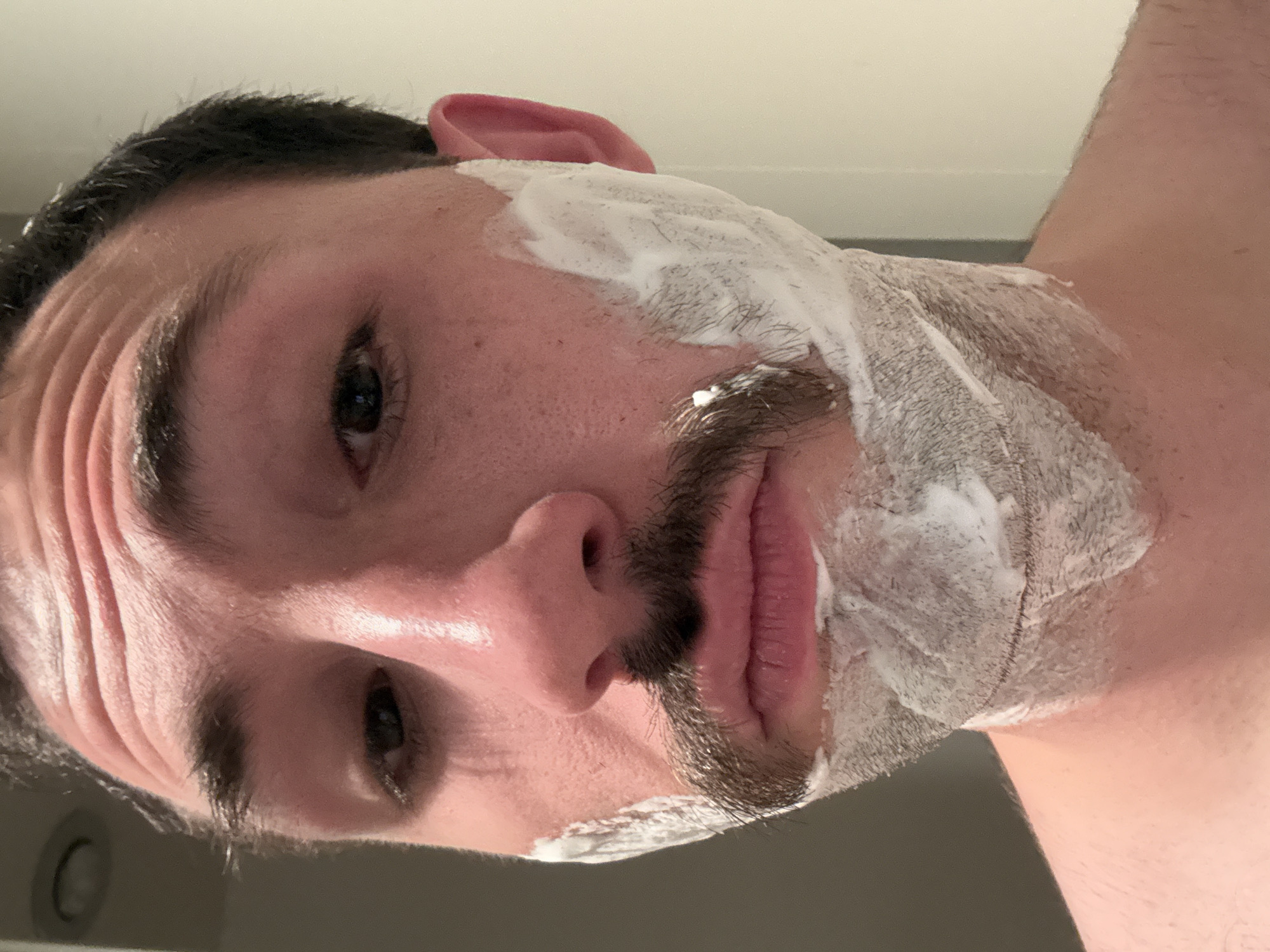 A low-angle, close-up portrait captures a person, likely male, engaged in a shaving routine. The image is tilted, presenting the individual's face from a unique perspective, looking upwards towards the camera. White shaving cream generously covers the jawline, cheeks, and upper neck area, extending partially onto the right ear. Dark stubble and hair are visible through and around the foam, forming a mustache above the lips and along the lower face.
The person has dark hair and eyebrows. Their left eye is partially visible, appearing to gaze directly at the viewer. Distinct wrinkles are present on the forehead, possibly indicating a facial expression or natural skin folds. The skin on the face appears moist, suggesting preparation for shaving. The background is uniformly light-colored and out of focus, consistent with an indoor setting, potentially a bathroom, illuminated by bright overhead lighting. A portion of the person's bare upper body or shoulder is visible on the right side of the frame. No text is visible in the image.