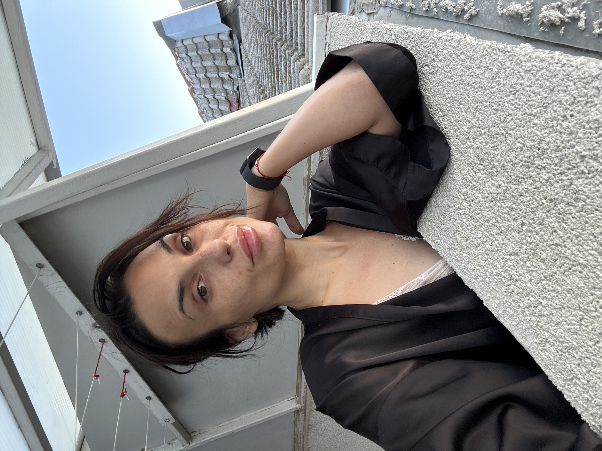 A woman with dark hair and freckles takes a selfie outdoors. She is wearing a black robe over a white lace bra. Her left arm is propped on a textured surface, and she has a smartwatch on her left wrist. Behind her, a clear blue sky is visible above a tiled roof. The location is the city of Pleven, Bulgaria.