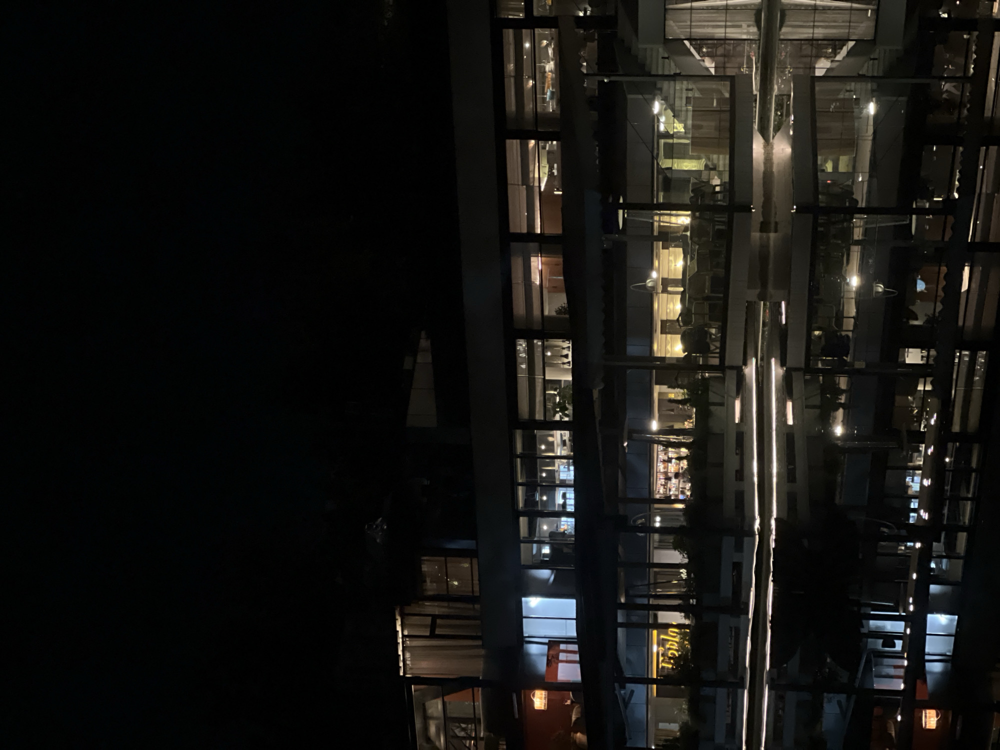 This night photograph, taken in Yerevan, Armenia, depicts a modern, multi-story building with a prominent glass facade. The image is dominated by a large, dark expanse on the left, indicating the night sky or an unlit area, while the building occupies the right side of the frame.
The building's numerous windows are illuminated from within, revealing glimpses into its interior spaces. These lights suggest that the building is active, likely housing commercial establishments such as a hotel, offices, or a restaurant/bar. On some lower floors, a bar area is visible through the windows, complete with bottles and glasses arranged on shelves. A partially obscured yellow sign can be seen in one of these windows, displaying text that appears to spell "Baplu" or "Bar."
Bright vertical lines of light are prominently reflected on the building's glass surfaces, creating a striking visual effect. These reflections likely originate from interior light fixtures or distinct architectural elements within the structure. The overall impression is one of a vibrant, contemporary urban environment at night. No specific individuals are clearly visible within the illuminated spaces, contributing to a serene yet active atmosphere. The image captures the modern design of the building and its illuminated presence against the dark backdrop of the night.